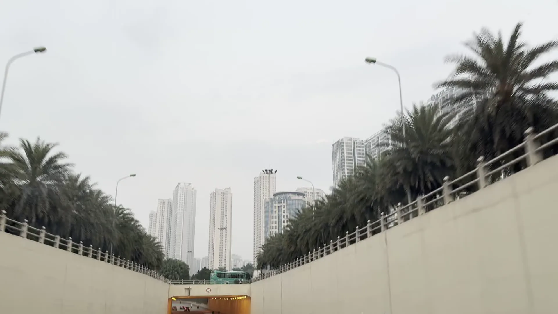 Đây là cảnh quay từ bên trong một chiếc ô tô đang di chuyển trên một con đường đô thị và đi qua một hầm chui. Các phương tiện khác như ô tô con và một chiếc xe buýt màu xanh lá cây cũng đang lưu thông trên đường. Hai bên hầm chui là những bức tường bê tông cao. Ở phía trên và trong tầm nhìn xa là nhiều tòa nhà chung cư cao tầng hiện đại. Dọc theo lối vào hầm chui có trồng nhiều cây cọ. Bầu trời u ám, và mặt đường có vẻ hơi ướt, cho thấy một ngày nhiều mây hoặc sau cơn mưa.