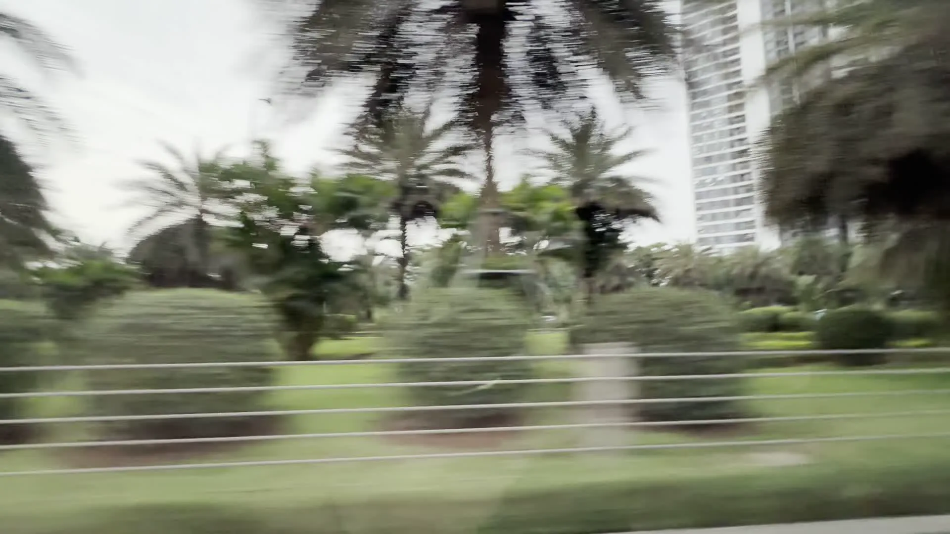 Quang cảnh được ghi lại từ một phương tiện đang di chuyển, nhìn ra một khu đô thị hiện đại. Ở phía sau là rất nhiều tòa nhà chung cư cao tầng màu trắng, san sát nhau. Dọc theo con đường là một khu vực cây xanh được chăm sóc cẩn thận, bao gồm nhiều cây cọ và các bụi cây được cắt tỉa thành hình tròn. Bầu trời có mây và màu xám. Một hàng rào chắn bằng kim loại chạy dọc ven đường ở tiền cảnh. Có một biển báo màu trắng nhưng chữ bị mờ do chuyển động nên không thể đọc được.