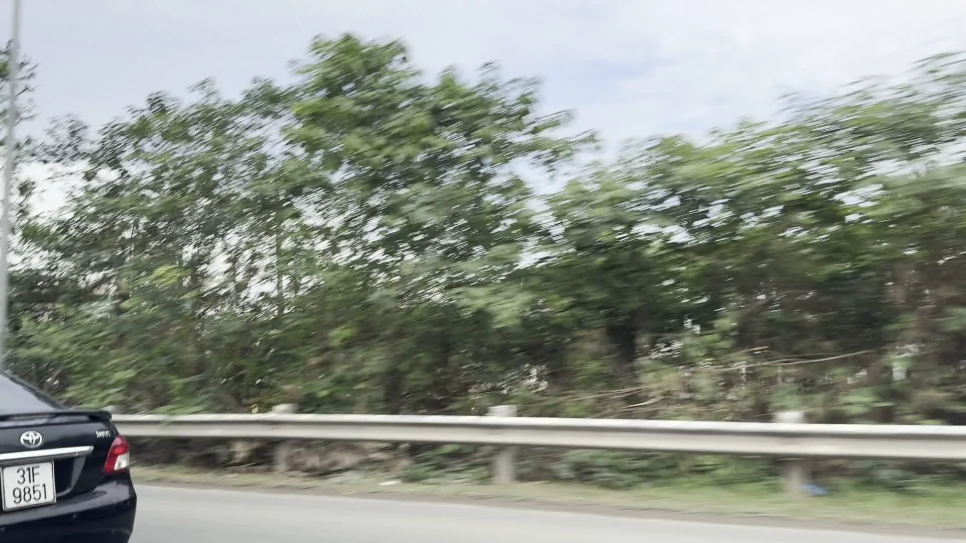 Hình ảnh được ghi lại từ một chiếc xe đang di chuyển, cho thấy quang cảnh bên đường. Phía trước có một chiếc ô tô màu đen đang chạy cùng chiều. Dọc theo con đường là một hàng rào chắn an toàn bằng kim loại. Phía sau hàng rào là cây cối rậm rạp. Bầu trời có nhiều mây. Qua các lùm cây, có thể nhìn thấy một công trình kiến trúc cao tầng đang trong quá trình xây dựng, có hình dáng giống một ngôi chùa hoặc đình.