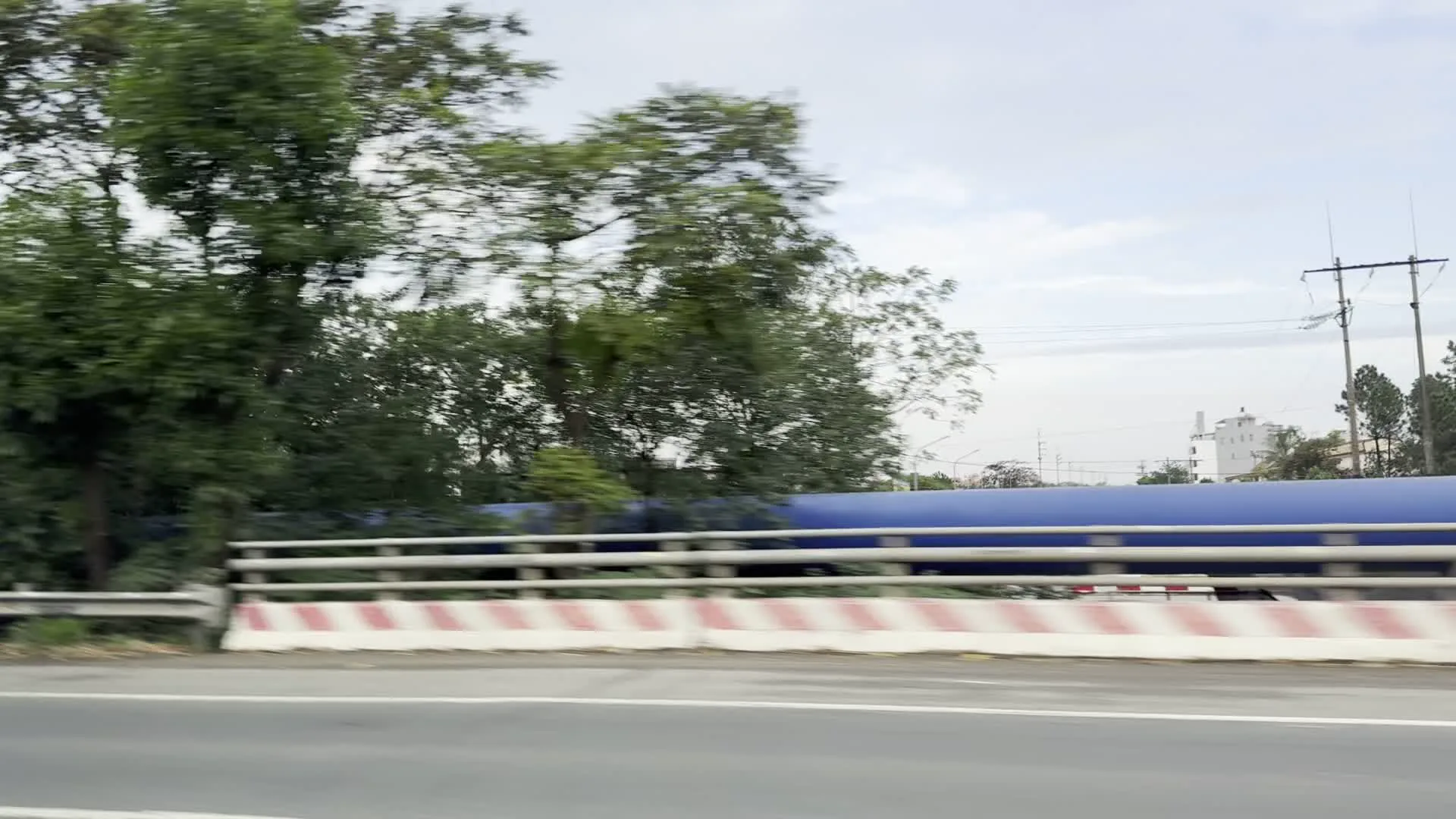Hình ảnh được chụp từ một phương tiện đang di chuyển, cho thấy quang cảnh ven đường trên một con đường quốc lộ. Con đường được trải nhựa, có lan can kim loại và một dải phân cách bằng bê tông sơn sọc đỏ trắng. Hai bên đường là thảm thực vật xanh tốt với nhiều cây cối rậm rạp, bao gồm cả cây chuối. Trong ảnh cũng có thể thấy một đường ống lớn màu xanh dương chạy song song với đường, các cột điện và một tòa nhà màu trắng ở phía xa. Do xe đang chạy nên khung cảnh có phần bị mờ. Không có hoạt động cụ thể nào của con người được ghi lại ngoài việc di chuyển trên đường.