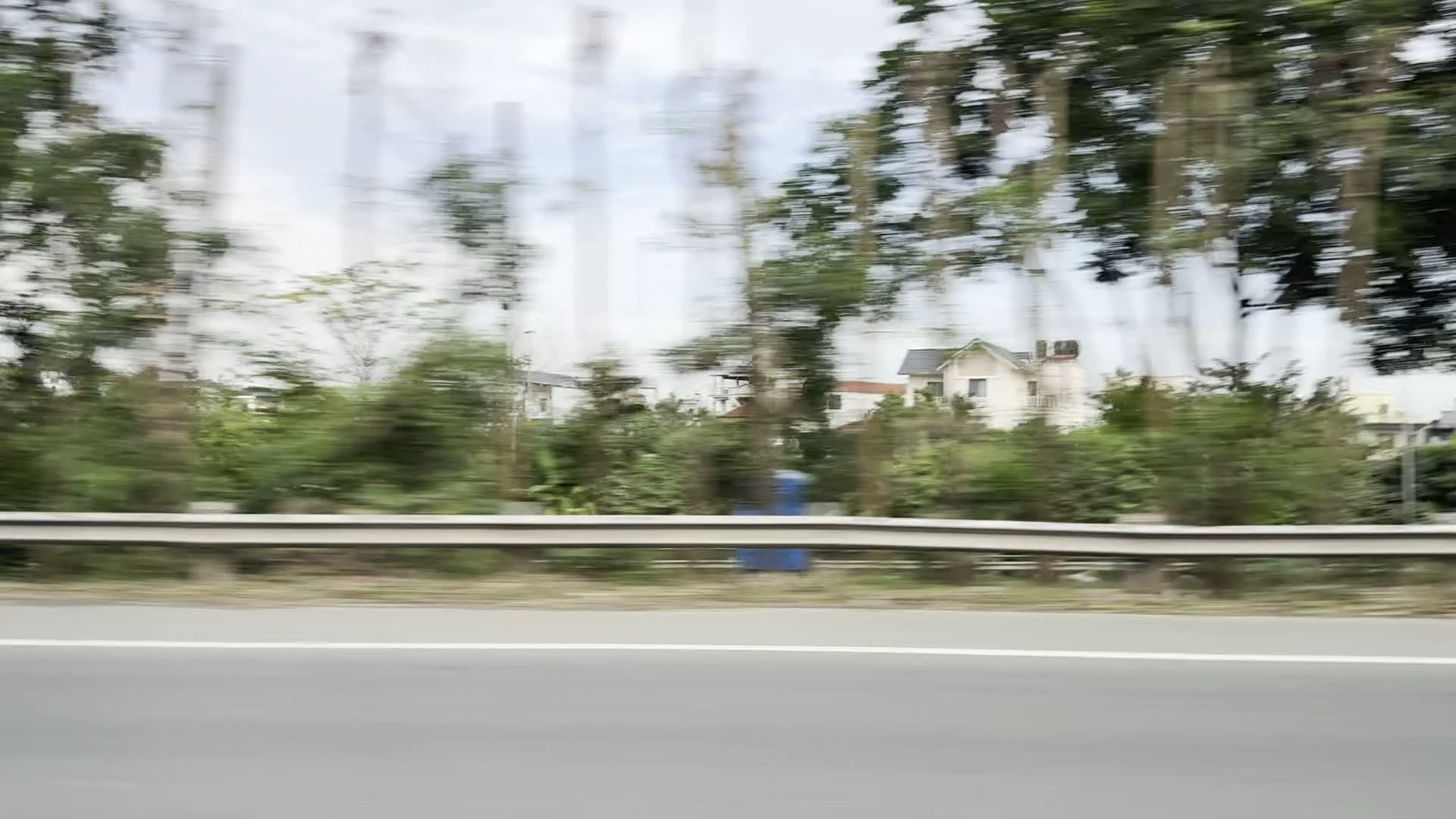 Cảnh quan được ghi lại từ một phương tiện đang di chuyển với tốc độ cao trên một con đường lớn hoặc đường cao tốc, điều này thể hiện qua các hình ảnh bị nhòe do chuyển động. Bên lề đường có một dải lan can bảo vệ bằng kim loại. Hai bên đường là cảnh quan kết hợp giữa khu dân cư với các ngôi nhà, tòa nhà và cây cối xanh um tùm. Một chiếc xe tải phủ bạt đang di chuyển cùng chiều. Có một biển báo giao thông hình tam giác nền vàng nhưng không thể nhận dạng được ký hiệu bên trong.