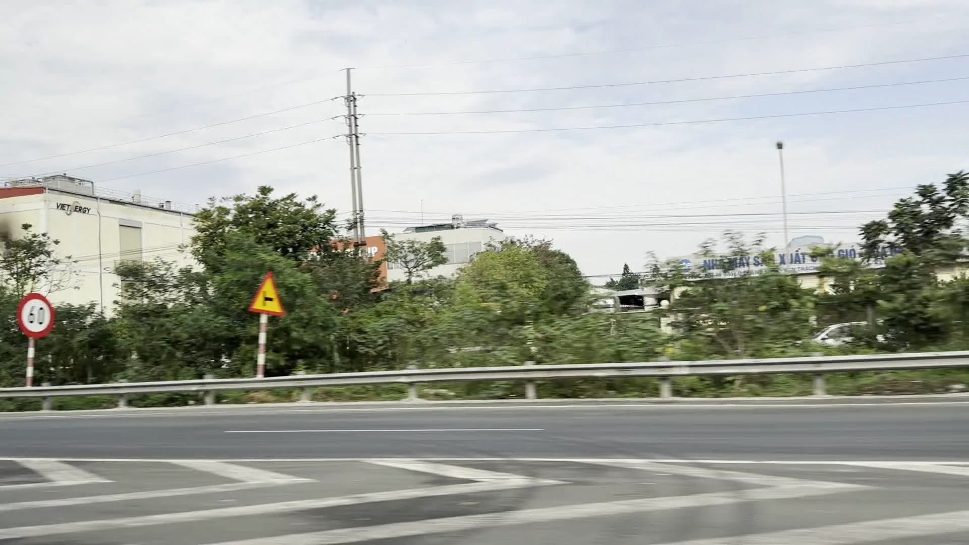 Hình ảnh được chụp từ một phương tiện đang di chuyển ven một con đường quốc lộ hoặc đường cao tốc. Cảnh vật bao gồm:。- Một con đường nhựa có vạch kẻ đường hình chữ V màu trắng.。- Hàng rào chắn bằng kim loại chạy dọc ven đường.。- Cây cối và bụi rậm mọc um tùm bên đường.。- Biển báo giao thông: một biển báo giới hạn tốc độ hình tròn, viền đỏ, nền trắng với số "60" ở giữa; và một biển báo nguy hiểm hình tam giác màu vàng nhưng không rõ biểu tượng bên trong.。- Ở phía sau, có một tòa nhà màu trắng với dòng chữ "VIETERGY". Một tòa nhà khác có vẻ là nhà máy với dòng chữ màu xanh lam, một phần có thể đọc được là "...NHÀ MÁY SẢN XUẤT GIẤY...".。- Một cột điện cao thế và dây điện chằng chịt.。Các hình ảnh khác cho thấy cảnh vật ven đường bị nhòe đi do tốc độ di chuyển nhanh của phương tiện, chủ yếu là hình ảnh hàng cây lướt qua.