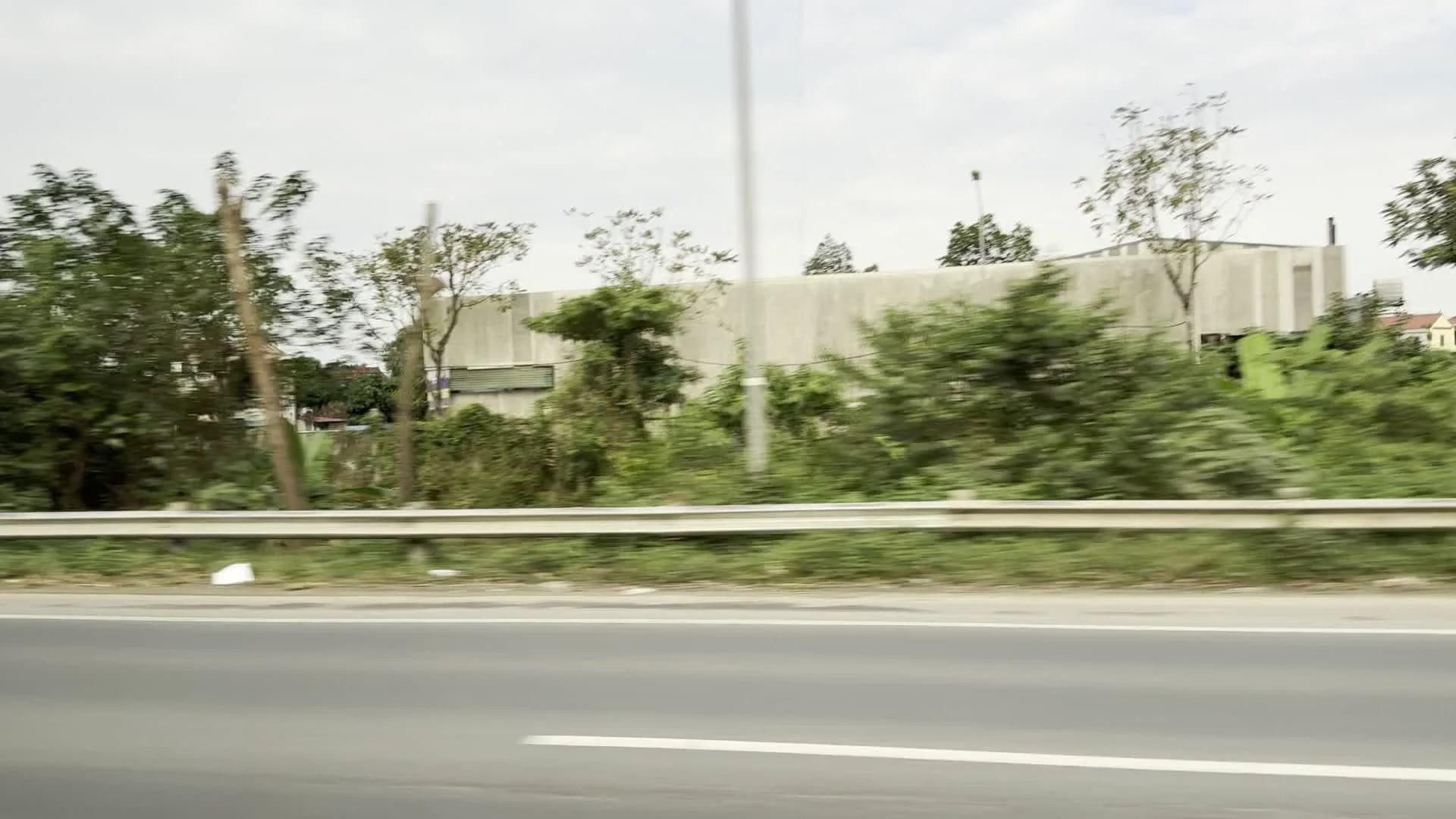 Đây là chuỗi hình ảnh được chụp từ một phương tiện đang di chuyển, cho thấy cảnh quan ven một con đường nhựa có lan can an toàn bằng kim loại. Bên kia lan can là một khu vực có nhiều cây xanh và bụi rậm. Xa hơn phía sau, có thể nhìn thấy các công trình xây dựng và nhà dân, bao gồm một tòa nhà bê tông lớn có vẻ còn mới hoặc đang xây dựng, và những ngôi nhà khác thấp thoáng sau rặng cây. Do xe đang chạy với tốc độ cao nên hình ảnh bị mờ do chuyển động. Không có hoạt động cụ thể nào của con người được nhìn thấy. Không có biển báo hay chữ viết nào có thể đọc được.