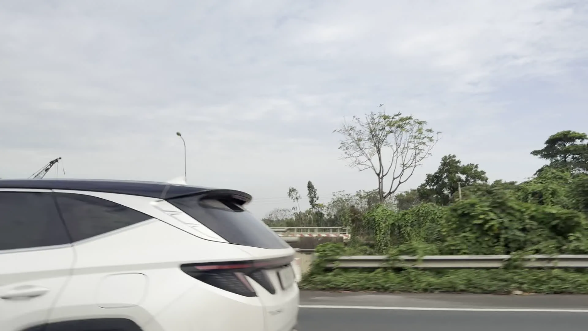 Quang cảnh được ghi lại từ một chiếc xe đang di chuyển trên đường cao tốc hoặc đường quốc lộ. Hình ảnh cho thấy một chiếc ô tô SUV màu trắng đang chạy cùng chiều. Hai bên đường có lan can bảo vệ bằng kim loại và dải phân cách bê tông sơn sọc đỏ trắng. Xung quanh là cây cối và thảm thực vật xanh tốt. Ở phía xa có thể thấy một cây cầu vượt và một phần của cần cẩu xây dựng. Bầu trời nhiều mây. Một số hình ảnh bị mờ do xe di chuyển với tốc độ cao. Không có văn bản hay biển hiệu nào có thể đọc được trong các hình ảnh.