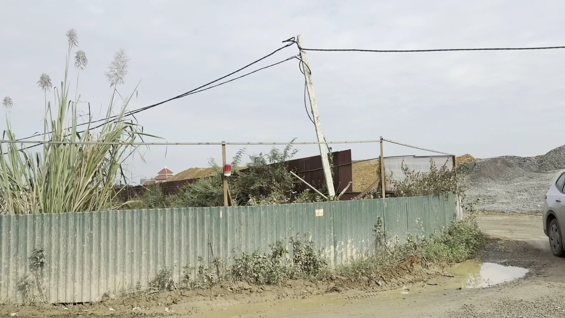 Khung cảnh cho thấy một khu vực có vẻ là bãi chứa vật liệu xây dựng, được bao quanh bởi một hàng rào làm bằng tôn lượn sóng màu xanh đã cũ. Bên trong hàng rào, có thể thấy các đống sỏi và cát lớn. Một cột điện bê tông và dây điện chạy ngang qua phía trên. Phía trước hàng rào, mặt đất lầy lội, có một vũng nước và cỏ dại mọc um tùm, bao gồm cả những cây lau sậy cao đang trổ bông. Ở rìa phải của hình ảnh đầu tiên, có thể thấy một phần của một chiếc ô tô màu bạc. Phía xa xa sau hàng rào có thể nhìn thấy một phần mái nhà.
