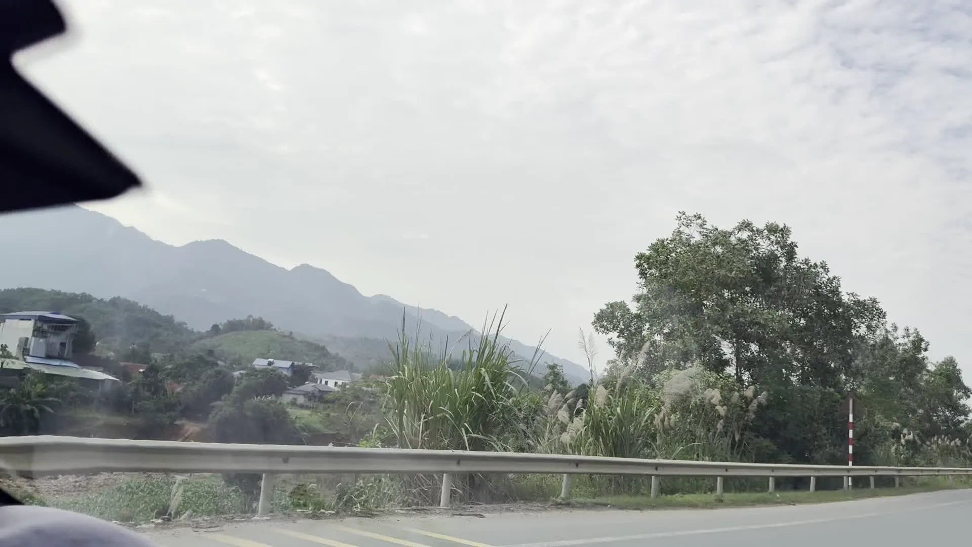 Cảnh tượng được ghi lại từ bên trong một chiếc ô tô đang di chuyển trên một con đường nhựa. Khung cảnh bên ngoài là một vùng nông thôn miền núi. Ở phía trước, có thể thấy một phần của chiếc xe. Ven đường là một hàng rào chắn bằng kim loại chạy dọc theo con đường. Xa hơn là những sườn đồi với cây cối xanh tươi và một vài ngôi nhà nằm rải rác. Phía sau cùng là những dãy núi cao trập trùng, bao phủ bởi một lớp sương mờ. Bầu trời có nhiều mây trắng, thời tiết có vẻ âm u.