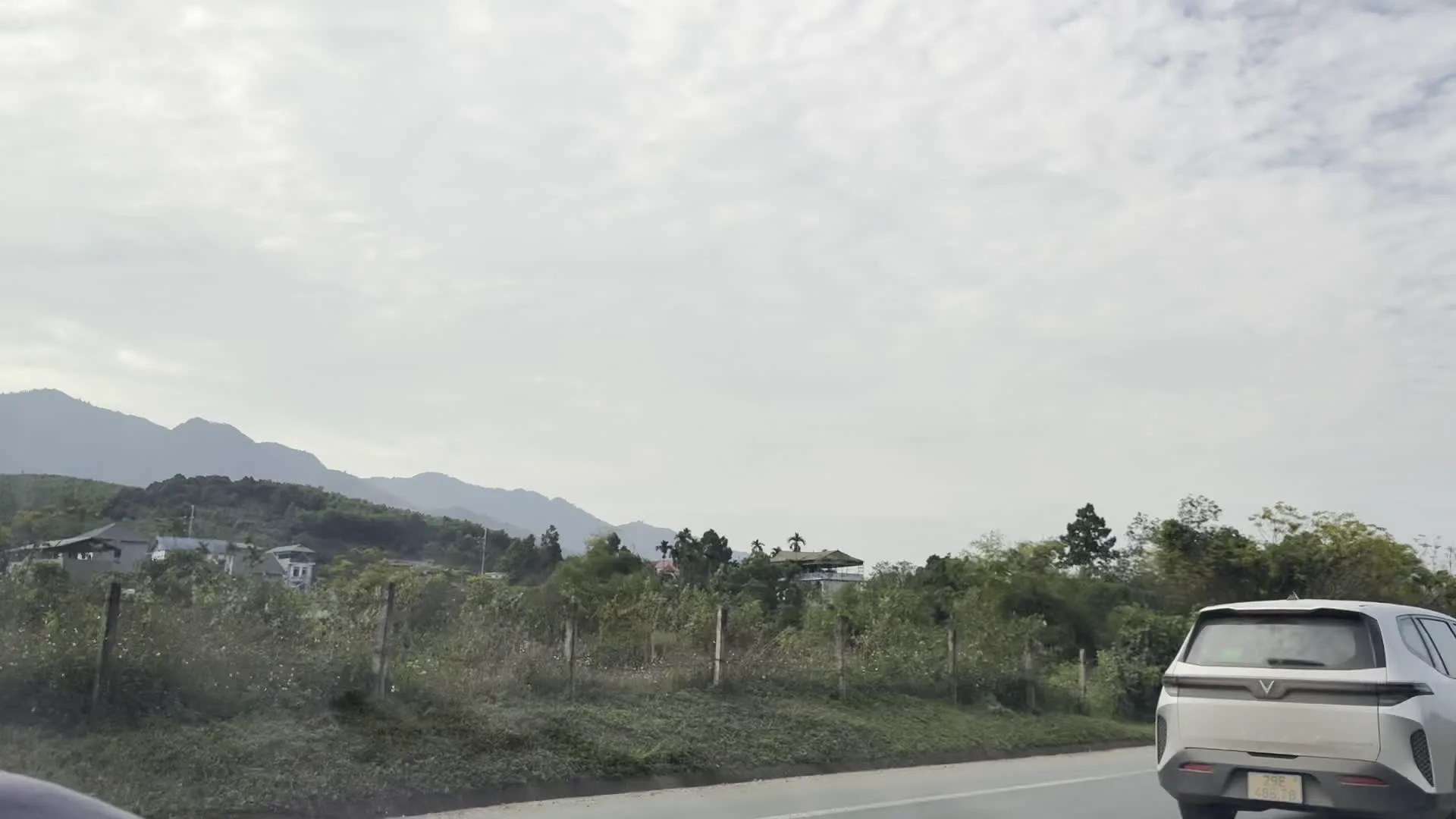 Cảnh quay từ bên trong một chiếc ô tô đang di chuyển trên một con đường quốc lộ hoặc đường cao tốc ở khu vực nông thôn, miền núi. Phong cảnh hai bên đường có nhiều cây cối xanh tươi, đồi núi trập trùng ở phía xa. Có một chiếc ô tô màu trắng đang chạy phía trước cùng chiều. Dọc ven đường có các hàng rào chắn bảo vệ. Bầu trời nhiều mây.