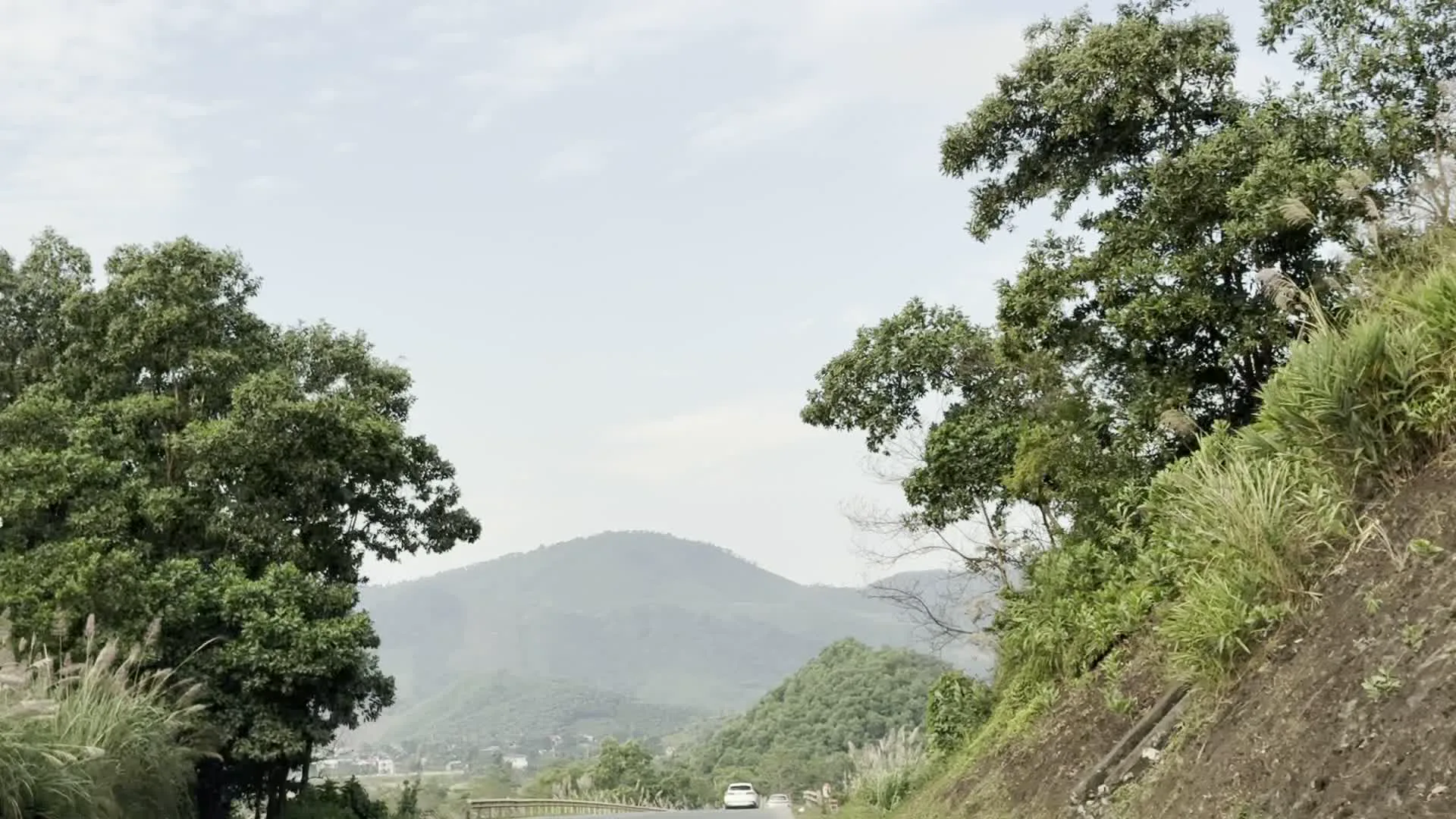 Cảnh quay ghi lại hành trình di chuyển trên một con đường ở vùng núi, có thể là khu vực Hòa Bình. Khung cảnh được nhìn từ bên trong một phương tiện đang chạy, có thể là ô tô. Con đường có lan can bảo vệ bằng kim loại. Hai bên đường là cây cối xanh tốt, rậm rạp, bao gồm cả cây lớn và những bụi cỏ lau. Phía xa là những dãy núi trập trùng được bao phủ bởi rừng cây. Trên đường có một chiếc ô tô màu trắng đang di chuyển. Bầu trời có mây, thời tiết quang đãng.