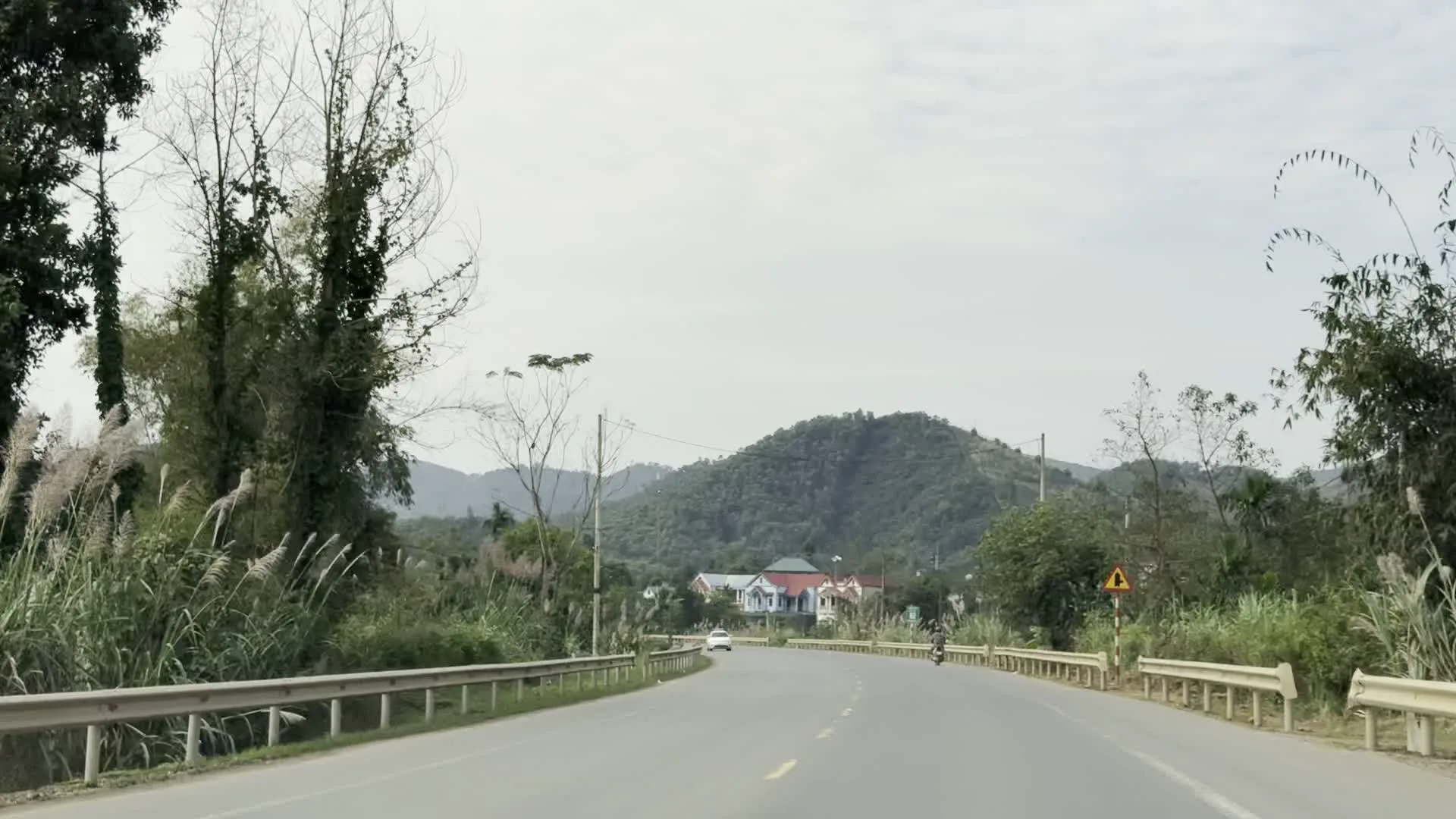 Quang cảnh được ghi lại từ một chiếc xe đang di chuyển trên một con đường nhựa ở khu vực nông thôn, miền núi. Hai bên đường là lan can bảo vệ bằng kim loại và cỏ lau mọc um tùm. Phía trước có một chiếc ô tô màu trắng đang chạy trên đường và một biển báo giao thông hình tam giác màu vàng, nhưng không thể nhìn rõ ký hiệu bên trong. Xa xa là những dãy núi trập trùng và một vài ngôi nhà dân nằm rải rác dưới chân núi.