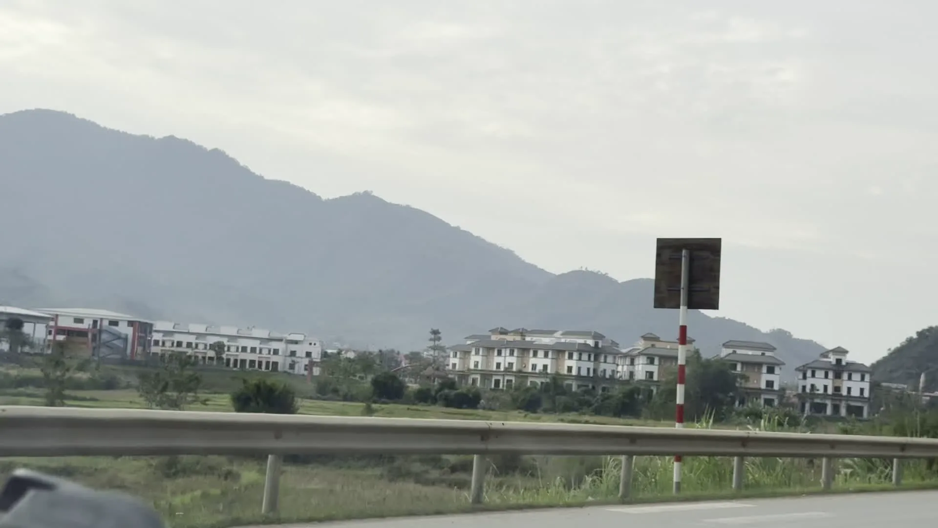 Đây là một loạt hình ảnh được chụp từ một chiếc xe đang di chuyển trên một con đường ở khu vực miền núi, có thể là tỉnh Hòa Bình. Phong cảnh chủ đạo là những dãy núi trập trùng, mờ ảo trong sương dưới bầu trời u ám. Dọc hai bên đường có thể thấy lan can an toàn bằng kim loại, cây cối và những bụi cỏ lau. Có những tòa nhà nhiều tầng với kiến trúc hiện đại, trông giống như một khu dân cư hoặc biệt thự mới xây. Trên đường có các phương tiện giao thông khác như ô tô và một chiếc xe buýt màu xanh lam đang đỗ bên đường. Trên thân xe buýt có dòng chữ "ĐIỆN ÔTÔ" và các thông tin quảng cáo dịch vụ sửa chữa. Ngoài ra còn có các biển báo giao thông, bao gồm một biển báo hiệu có trạm xăng ở phía trước. Một tòa nhà ven đường có treo biển quảng cáo lớn nhưng khó đọc, có thể nhận ra một vài từ liên quan đến dịch vụ sơn như "SƠN SẮT - INOX", "BÁN BUÔN BÁN LẺ CÁC LOẠI SƠN".
