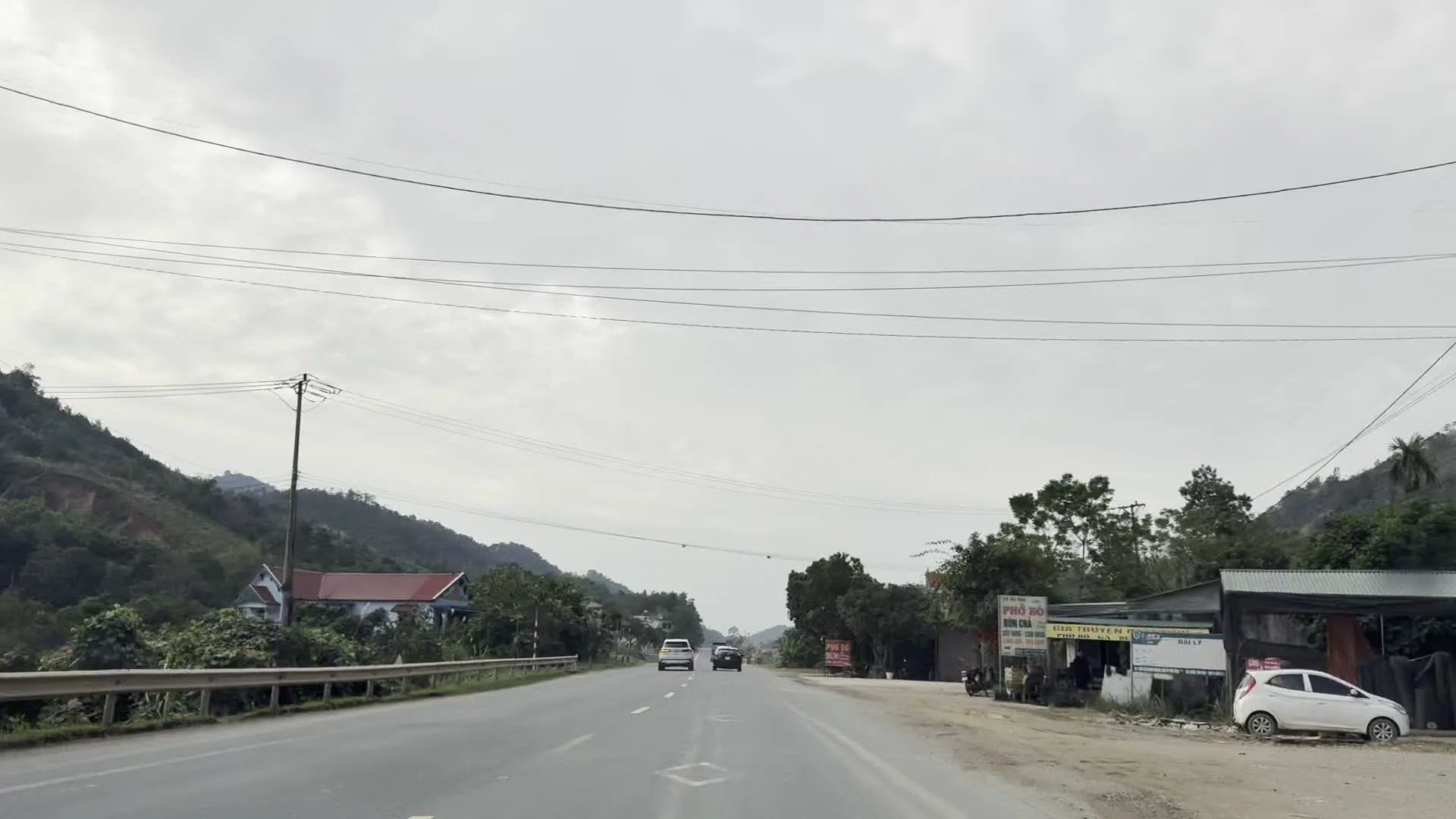 Đây là một loạt hình ảnh ghi lại cảnh quan trên một con đường quốc lộ ở vùng đồi núi. Con đường trải nhựa rộng rãi với nhiều xe ô tô đang lưu thông. Hai bên đường là đồi núi trập trùng, cây cối và những bụi lau sậy nở hoa trắng.。Trong các bức ảnh có thể nhận thấy:。- Một quán ăn ven đường với biển hiệu có các chữ "PHỞ BÒ", "NƯỚC MÍA", và các chữ khác không rõ ràng.。- Một tấm biển quảng cáo lớn ở bên trái đường có logo của "SACOMBANK" và dòng chữ "VĨNH DÃ KỲ HOA".。- Một biển báo giao thông hình tam giác viền xanh, nền trắng cảnh báo đường không bằng phẳng.。- Một số ngôi nhà dân, bao gồm một ngôi nhà mái đỏ nằm trên sườn đồi.。Khung cảnh chung cho thấy một tuyến đường giao thông xuyên qua khu vực trung du hoặc miền núi phía Bắc Việt Nam, với bầu trời u ám, nhiều mây.