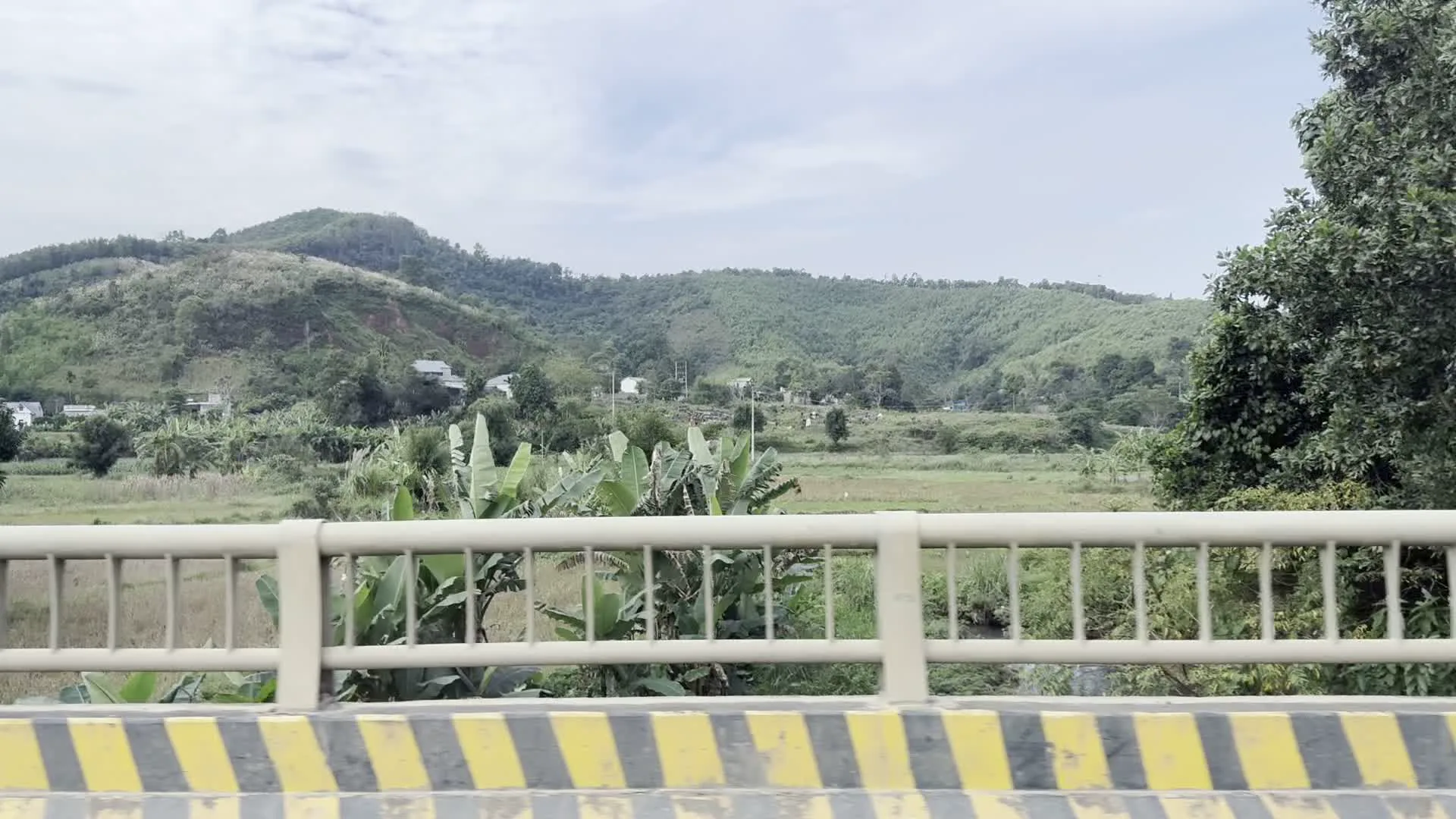 Cảnh quay từ một phương tiện đang di chuyển trên đường, ghi lại khung cảnh nông thôn miền núi. Phía xa là những dãy đồi núi trập trùng được bao phủ bởi cây xanh. Rải rác dưới chân đồi là những ngôi nhà dân. Ở gần hơn là các khu đất nông nghiệp như ruộng lúa và vườn chuối. Một phần của cây cầu với lan can màu be và lề đường sơn sọc vàng đen xuất hiện trong một khung hình. Các khung hình khác cho thấy lan can bảo vệ ven đường và một tảng đá lớn bên vệ đường với thảm thực vật um tùm.