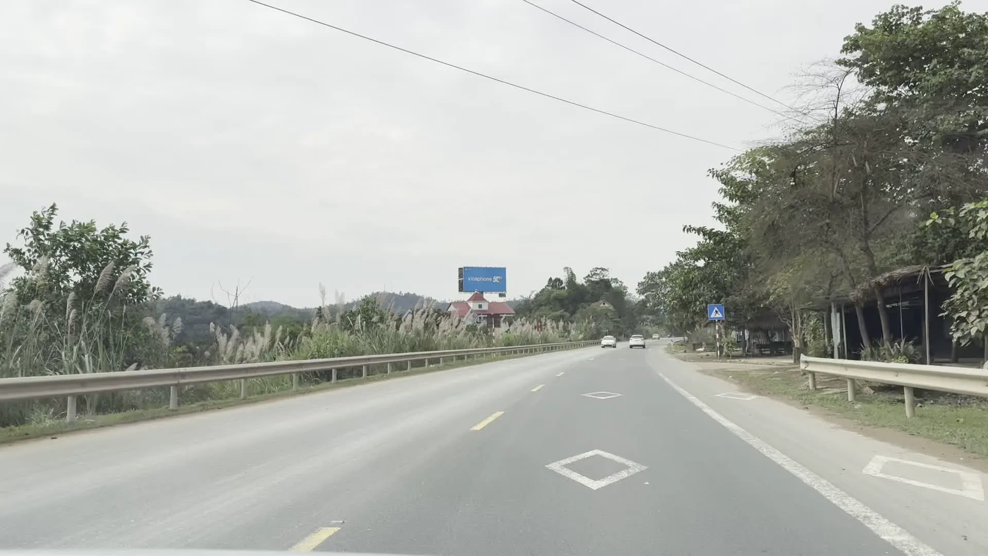 Hình ảnh ghi lại cảnh quan trên một con đường nhựa ở khu vực nông thôn hoặc miền núi. Bầu trời nhiều mây. Có một vài phương tiện như ô tô con và xe tải đang lưu thông trên đường. Hai bên đường là cây cối xanh tươi, cỏ lau và đồi núi. Bên đường có một biển quảng cáo lớn màu xanh của hãng lốp xe "BRIDGESTONE". Ngoài ra, có thể thấy một quán nhỏ ven đường, có thể là một tiệm sửa chữa lốp ô tô, với các biển hiệu ghi chữ "LỐP Ô TÔ" và "SỬA CHỮA", và một vài chiếc xe tải đang đỗ gần đó.