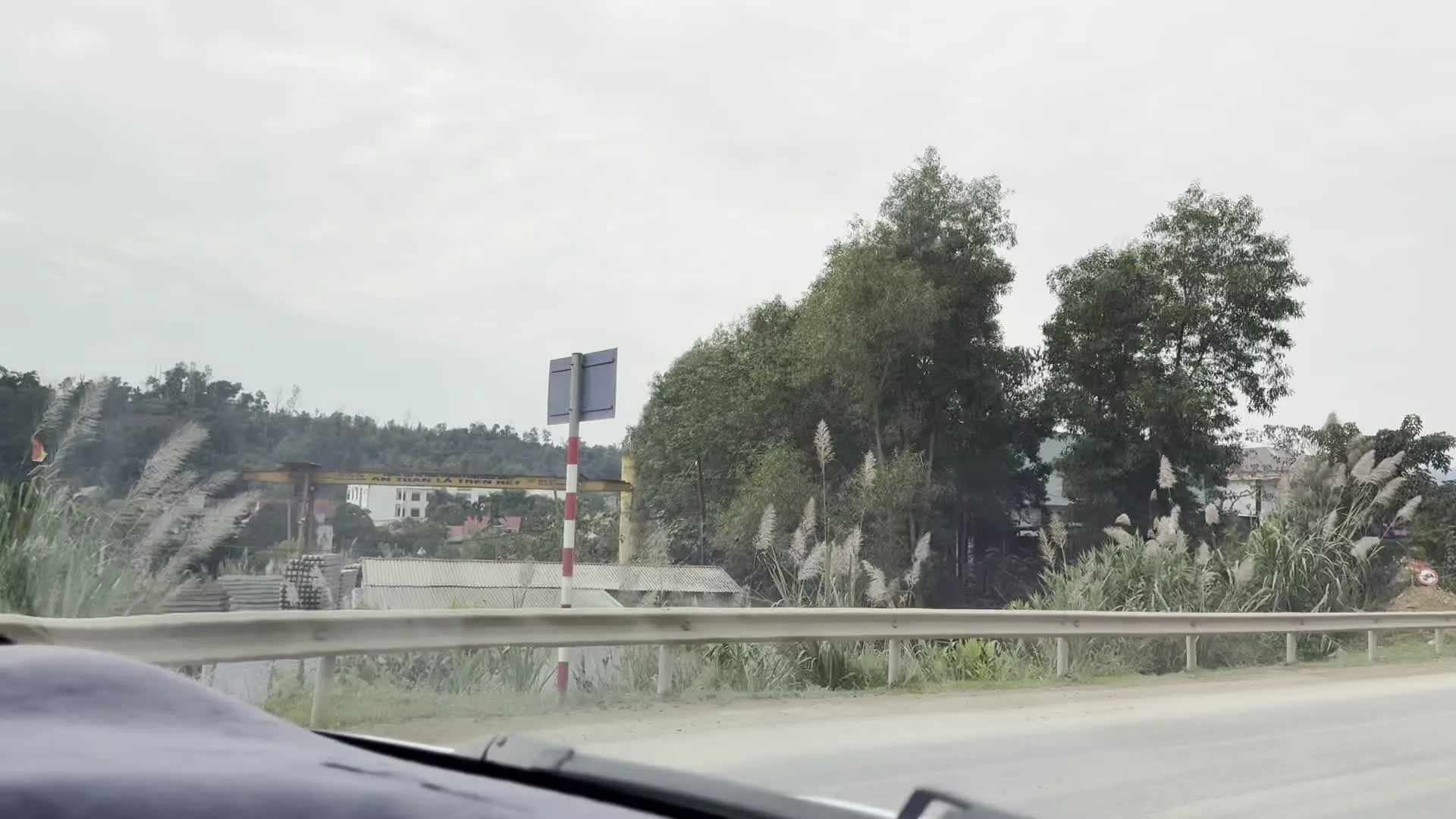 Đây là những hình ảnh được chụp từ bên trong một chiếc ô tô đang di chuyển trên một con đường ở khu vực nông thôn, miền núi. Phong cảnh xung quanh chủ yếu là cây cối xanh tươi và những dãy núi trập trùng phía xa dưới bầu trời nhiều mây. Con đường được trải nhựa, có lan can bảo vệ bằng kim loại hoặc bê tông ở hai bên. Có thể thấy một chiếc cần cẩu giàn lớn màu vàng, trên đó có khẩu hiệu "AN TOÀN LÀ TRÊN HẾT", và phía dưới là các vật liệu xây dựng như cống bê tông. Ngoài ra, trên đường còn có các biển báo giao thông hình tam giác nền vàng, viền đỏ, cảnh báo đoạn đường có người đi bộ cắt ngang. Một cột điện cao thế cũng xuất hiện ở ven đường.