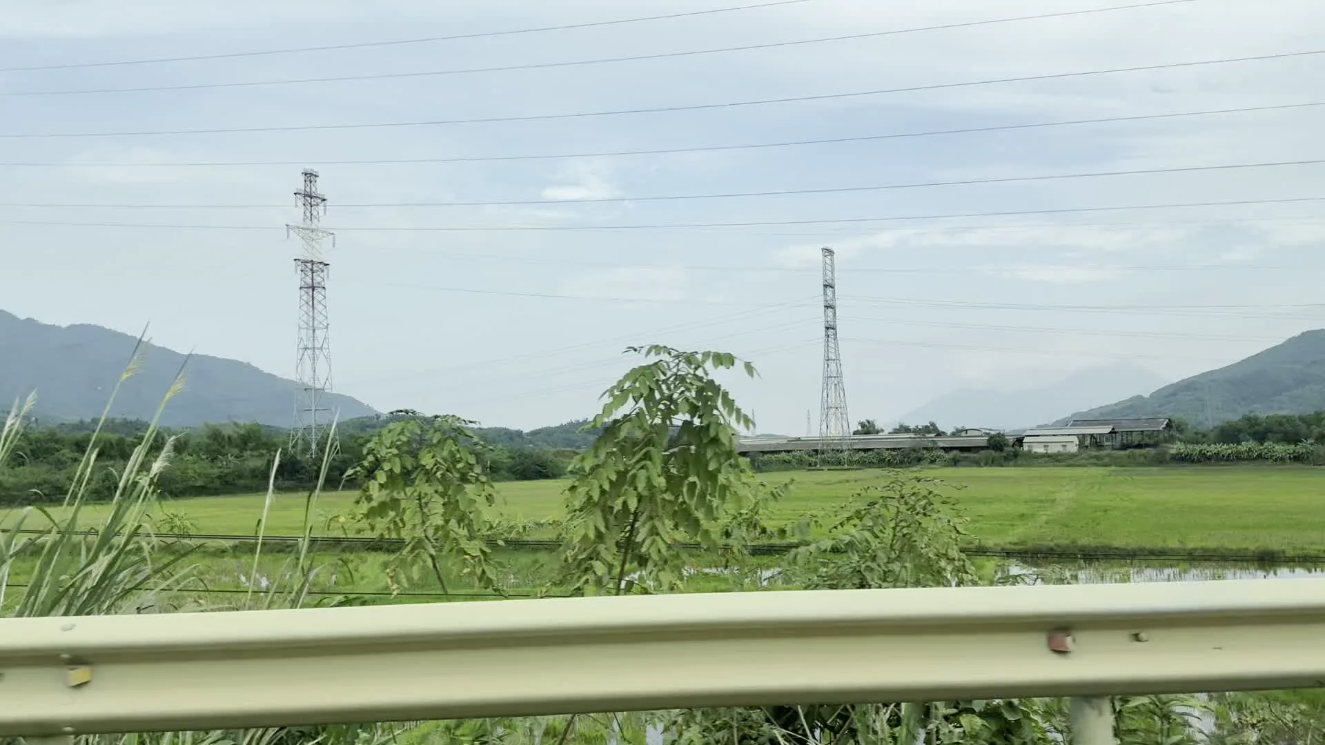 Quang cảnh vùng nông thôn miền núi được chụp từ một phương tiện đang di chuyển dọc theo con đường có lan can bảo vệ. Khung cảnh bao gồm những cánh đồng lúa xanh mướt, phía sau là những dãy núi trập trùng. Có hai cột điện cao thế bằng kim loại đứng sừng sững trên cánh đồng và một ngôi nhà mái ngói màu nâu đỏ nằm giữa cây cối xanh tươi trên sườn đồi. Bầu trời có nhiều mây, mang màu trắng xám. Không có người hay hoạt động cụ thể nào được nhìn thấy.