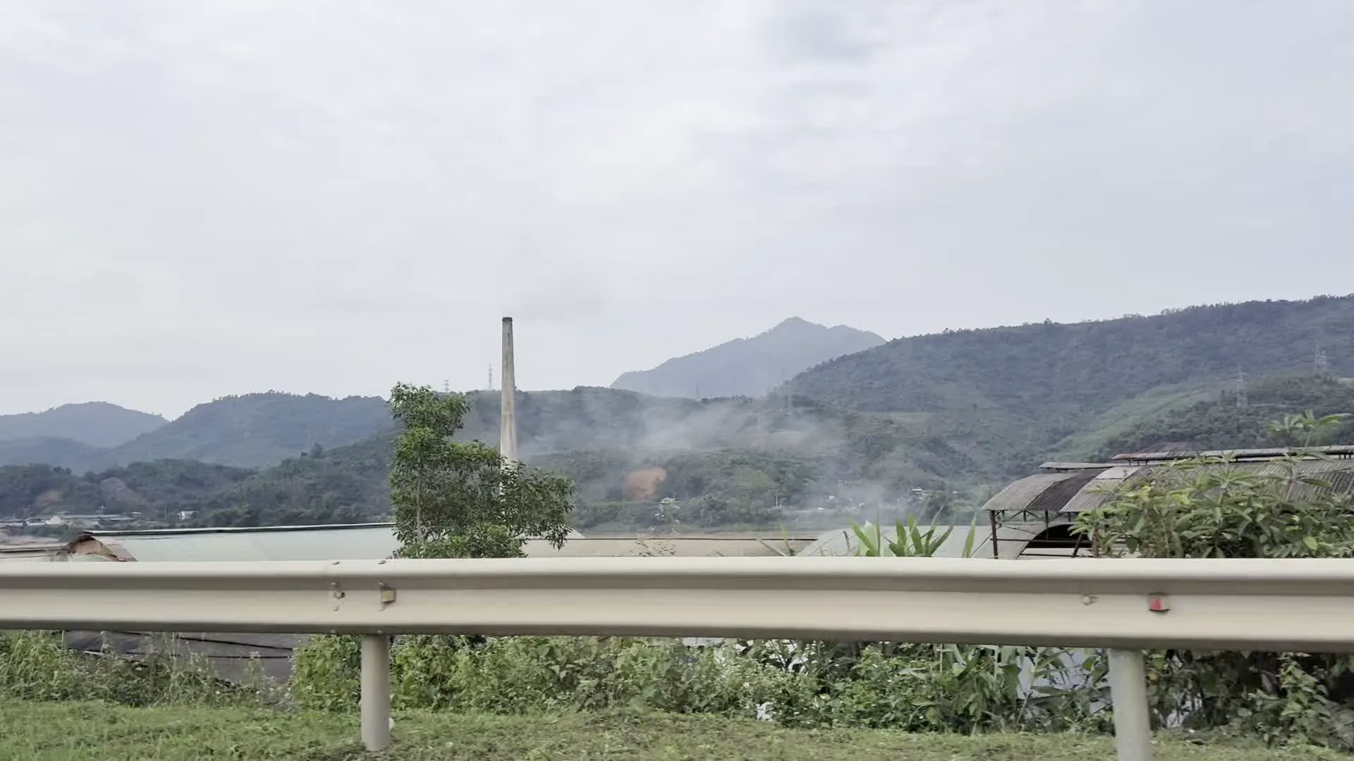 Hình ảnh ghi lại khung cảnh miền núi được chụp từ một phương tiện đang di chuyển trên đường hoặc cầu. Phía trước là lan can bảo vệ của đường bộ, có đoạn màu trắng, có đoạn sơn sọc vàng đen. Phong cảnh chính là những dãy núi trập trùng, bao phủ bởi cây xanh dưới bầu trời u ám. Nổi bật trong cảnh quan là một khu vực có vẻ là nhà máy hoặc cơ sở sản xuất công nghiệp, với một ống khói cao và làn khói trắng bốc lên. Xung quanh là các nhà xưởng có mái tôn, tường gạch và một vài ngôi nhà dân. Ngoài ra, còn có hình ảnh một con đường đất, cây cối, cột điện và một người đang đi bộ ở phía xa.