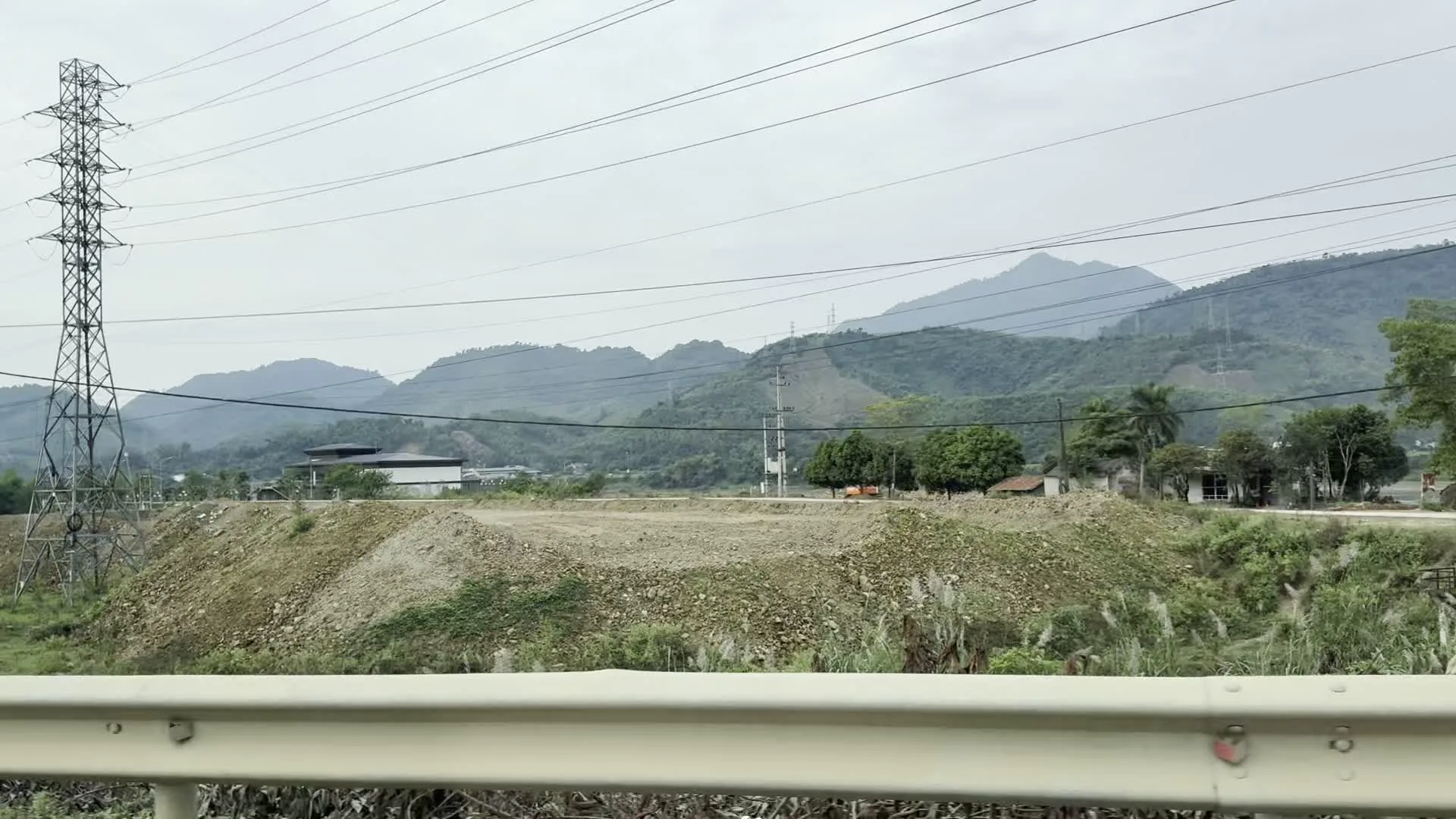 Bức ảnh chụp cảnh quan vùng nông thôn miền núi, có thể được chụp từ một phương tiện đang di chuyển trên đường có lan can bảo vệ. Nổi bật trong khung cảnh là một cột điện cao thế lớn bằng kim loại với hệ thống dây điện chằng chịt. Ở giữa là một khu đất rộng lớn đã được san lấp, với các ụ đất đá, cho thấy đây có thể là một công trường xây dựng hoặc khu vực khai thác. Phía sau là những dãy núi trập trùng cây cối bao phủ dưới bầu trời u ám. Rải rác có một vài ngôi nhà, cây xanh và một chiếc máy xúc màu cam thấp thoáng ở phía xa. Không có chữ viết rõ ràng nào có thể đọc được.