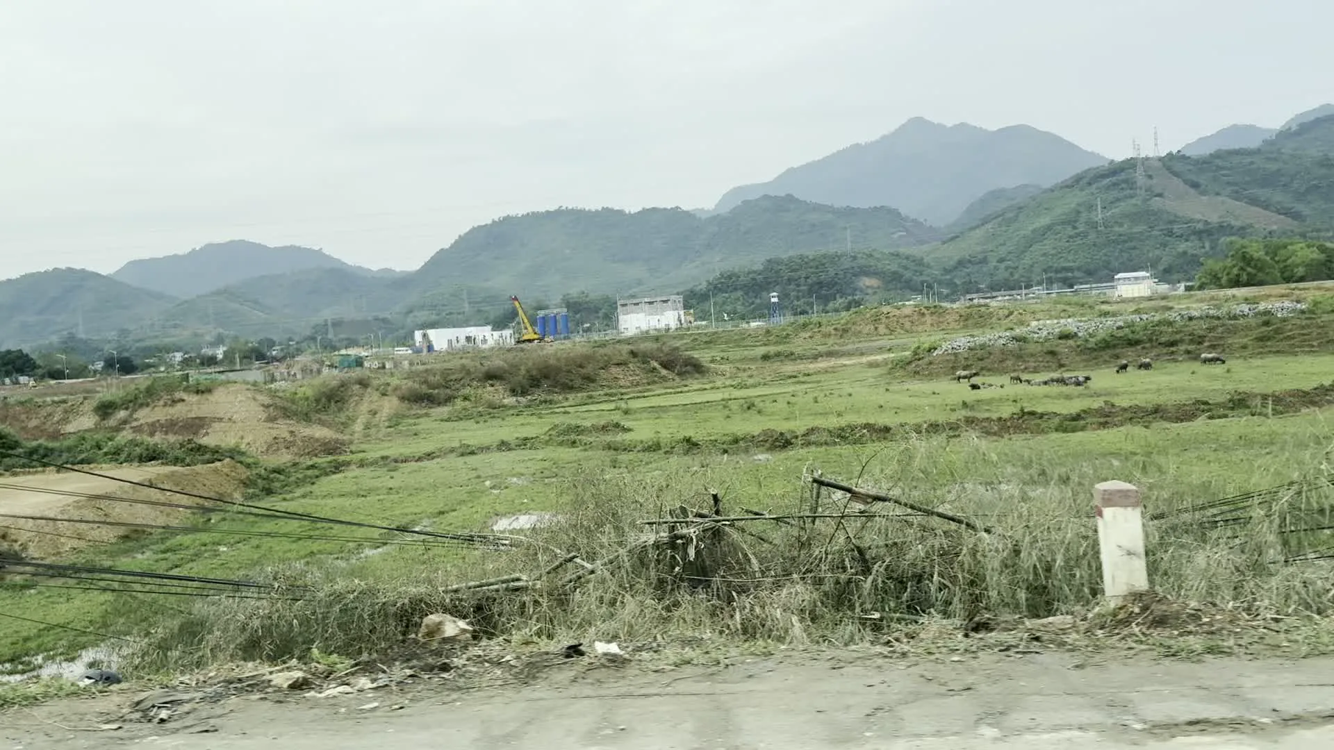 Hình ảnh ghi lại một quang cảnh vùng nông thôn miền núi. Ở hậu cảnh là những dãy núi trập trùng dưới bầu trời u ám, và có thể thấy các cột điện cao thế. Ở trung cảnh, có một khu vực đang được san lấp để xây dựng, với một chiếc cần cẩu màu vàng và một vài tòa nhà màu trắng. Bên cạnh khu công trường là một cánh đồng cỏ xanh, nơi một đàn trâu đang gặm cỏ. Tiền cảnh cho thấy ven một con đường có một cột mốc, cây cối mọc um tùm và nhiều dây điện, dây cáp lộn xộn. Không có văn bản hay chữ viết nào rõ ràng trong ảnh.