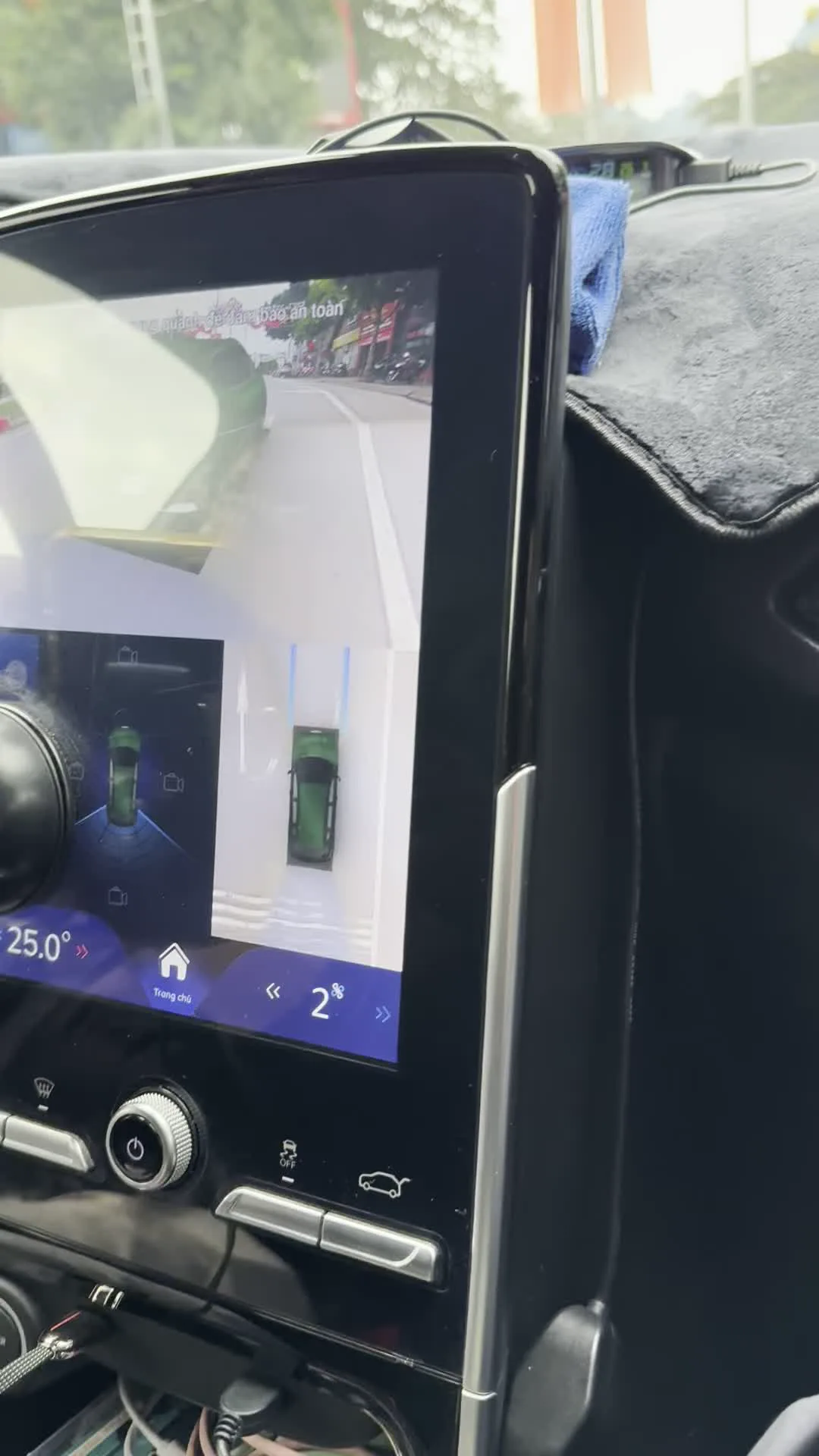 Bên trong một chiếc ô tô, hình ảnh tập trung vào màn hình điều khiển trung tâm đặt dọc. Màn hình này đang hiển thị hình ảnh từ hệ thống camera 360 độ, hỗ trợ người lái quan sát môi trường xung quanh xe. Trên màn hình có dòng chữ cảnh báo "Quan sát xung quanh để đảm bảo an toàn". Hệ thống đang chiếu hình ảnh một chiếc ô tô SUV màu xanh lá cây, có thể nhận ra là mẫu xe VinFast VF e34 qua logo và chữ ở đuôi xe. Giao diện màn hình hiển thị nhiệt độ "25.0°", nút "Trang chủ" và các điều khiển khác. Hoạt động đang diễn ra là người lái xe đang sử dụng hệ thống camera để di chuyển hoặc đỗ xe một cách an toàn trên đường phố.