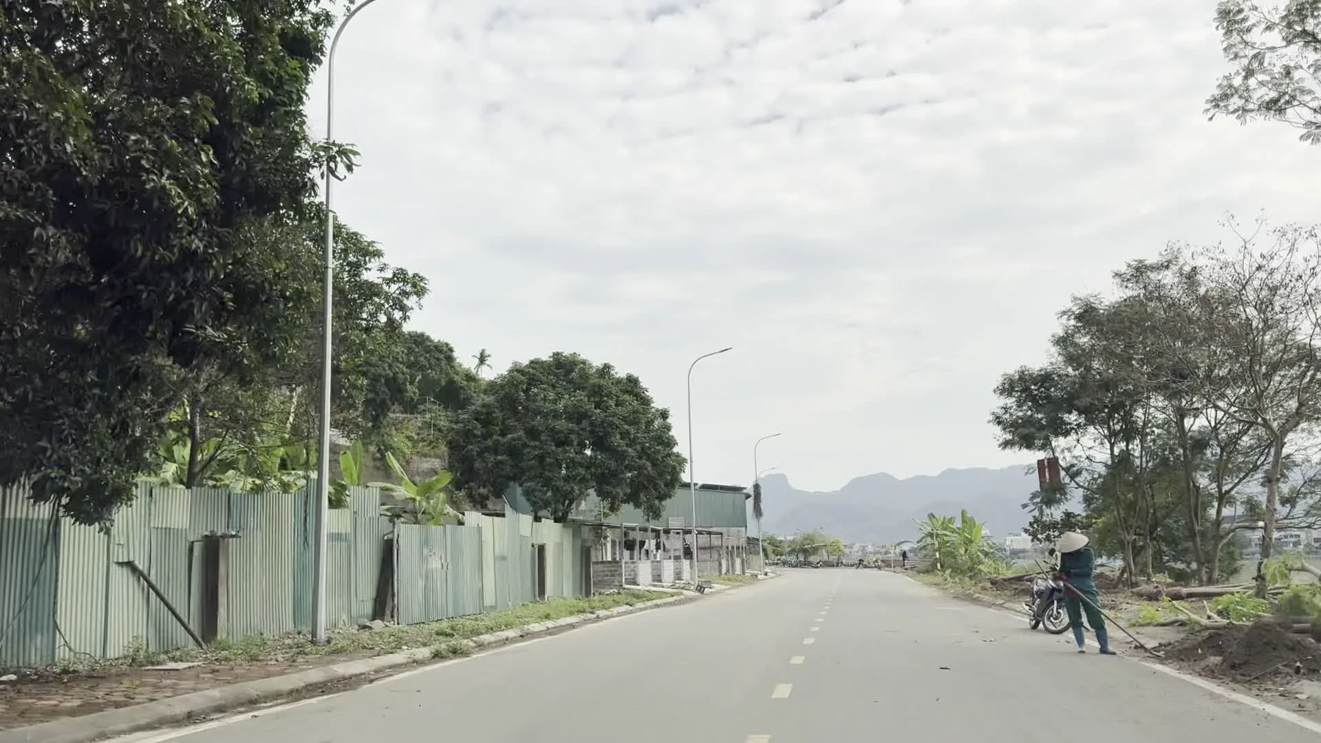 Hình ảnh chụp một con đường nhựa vắng vẻ ở khu vực nông thôn hoặc ngoại ô. Bầu trời nhiều mây, và ở phía xa có thể nhìn thấy những dãy núi. Dọc theo con đường có các cột đèn chiếu sáng. Bên trái là một hàng rào dài bằng tôn lượn sóng, phía sau có những ngôi nhà tạm và cây cối, bao gồm cả cây chuối. Bên phải đường, một người đội nón lá đang đứng làm việc, có thể đang quét dọn hoặc thu gom cành cây bên lề đường. Cạnh người đó là một chiếc xe máy đang đỗ. Không có chữ viết nào có thể đọc được trong hình.