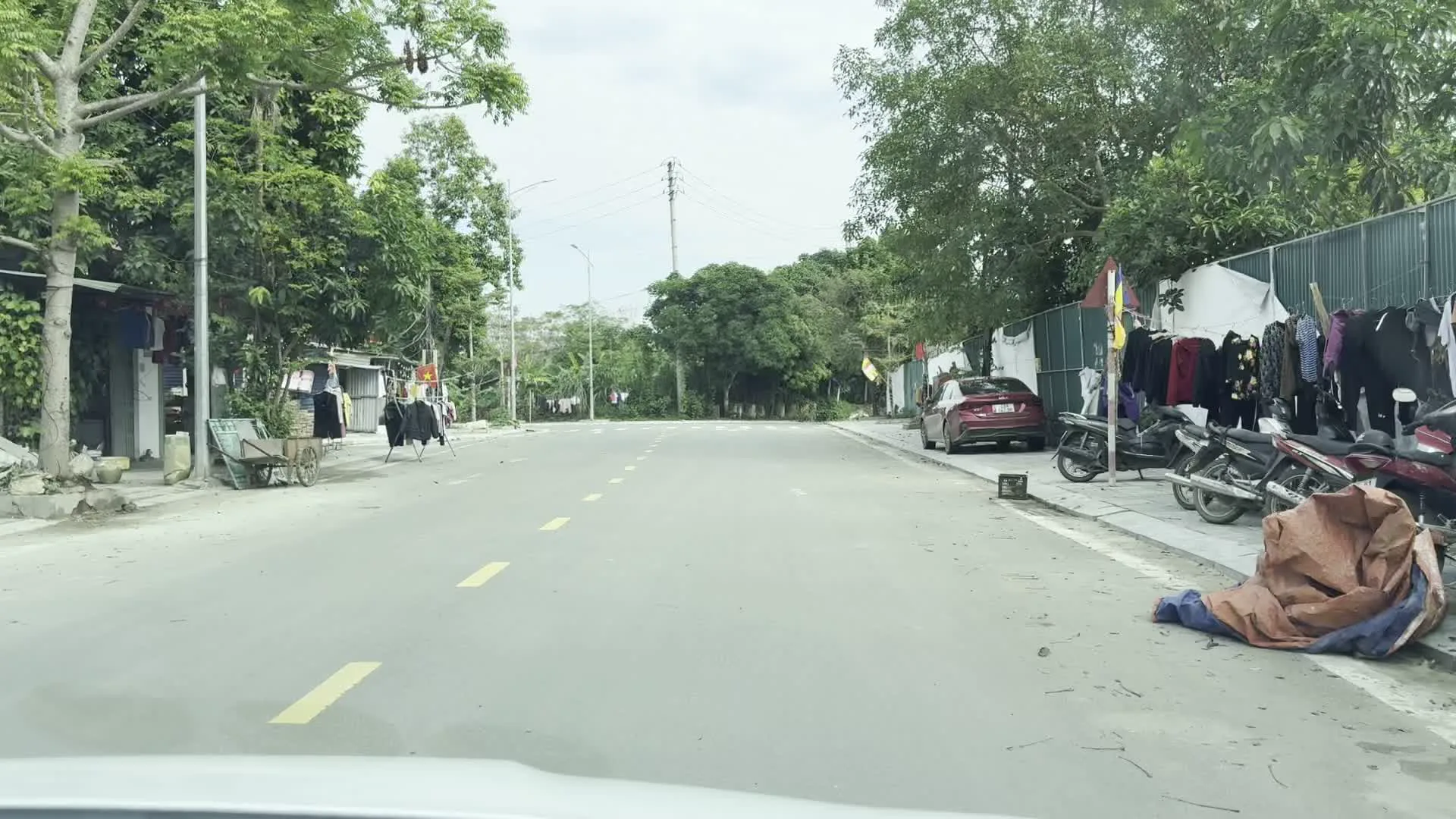 Đây là hình ảnh một con đường trải nhựa ở khu vực dân cư, có thể là ở vùng nông thôn hoặc ngoại ô tại tỉnh Hòa Bình. Con đường có vạch kẻ đường màu vàng đứt khúc.。Hai bên đường là cây cối xanh tươi, cột điện và nhà dân. Có thể thấy hoạt động sinh hoạt thường ngày của người dân. Cụ thể, dọc theo lề đường và vỉa hè, rất nhiều quần áo được treo trên các dây phơi hoặc giá treo, một số có vẻ như đang được bày bán. Một chiếc ô tô màu đỏ đang đỗ ở lề đường bên phải, cạnh một hàng rào tôn. Có người đi bộ trên vỉa hè và người điều khiển xe máy di chuyển trên đường. Một lá cờ đỏ sao vàng của Việt Nam được treo trên cột. Ở phía xa có thể nhìn thấy hình dáng của núi non, phù hợp với địa hình của khu vực này.