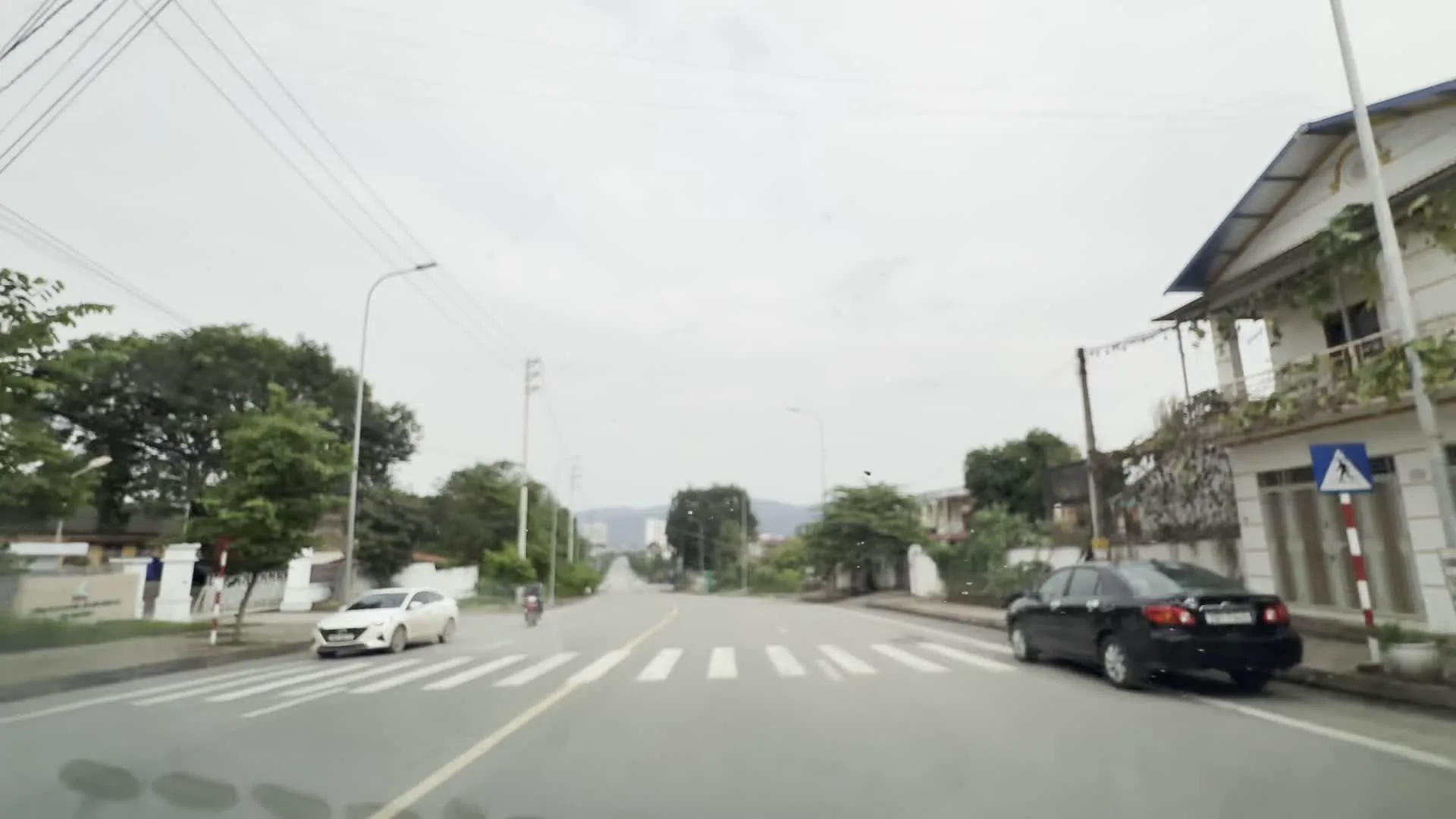 Cảnh quay được ghi lại từ một phương tiện đang di chuyển trên một con đường trải nhựa rộng rãi ở khu vực ngoại ô. Con đường có vạch kẻ phân làn, vạch cho người đi bộ qua đường và các mũi tên chỉ hướng. Hai bên đường là cây cối xanh tốt, nhà dân, cột điện và dây điện. Có một chiếc ô tô màu trắng đang di chuyển, một chiếc ô tô màu đen đang đỗ ven đường và một người đi xe máy ở phía xa. Hình ảnh cho thấy một ngã tư có đèn tín hiệu giao thông và các biển báo chỉ dẫn. Một ngôi nhà nhiều tầng sơn màu xanh lá cây nổi bật ở góc đường. Phía xa có thể thấy những dãy núi dưới bầu trời u ám.