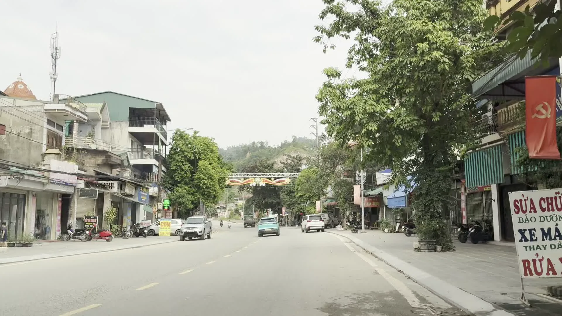 Đây là hình ảnh chụp một con đường tại một khu vực đô thị hoặc thị trấn ở Việt Nam, có thể là tại Hòa Bình. Con đường được trải nhựa và có nhiều phương tiện giao thông như ô tô và xe máy đang di chuyển. Hai bên đường là những dãy nhà cao tầng liền kề, đặc trưng của kiến trúc đô thị Việt Nam, với tầng trệt được sử dụng làm cửa hàng kinh doanh. Cây xanh được trồng dọc hai bên vỉa hè. Ở phía xa, có thể thấy một cổng chào lớn bắc ngang qua đường, được trang trí với hình ảnh Chủ tịch Hồ Chí Minh và các biểu ngữ màu đỏ chữ vàng. Có nhiều biển hiệu của các cửa hàng có thể đọc được, bao gồm: một biển hiệu lớn màu đỏ và trắng ghi "SỬA CHỮA - BẢO DƯỠNG - XE MÁY - THAY DẦU - RỬA XE"; một biển hiệu "DŨNG TRUỲN INOX NHÔM KÍNH"; một biển hiệu tiệm tóc "TÓC NAM". Ngoài ra, có một lá cờ Đảng Cộng sản Việt Nam (cờ búa liềm) treo trên một ngôi nhà. Khung cảnh tổng thể cho thấy một ngày sinh hoạt bình thường trên đường phố.
