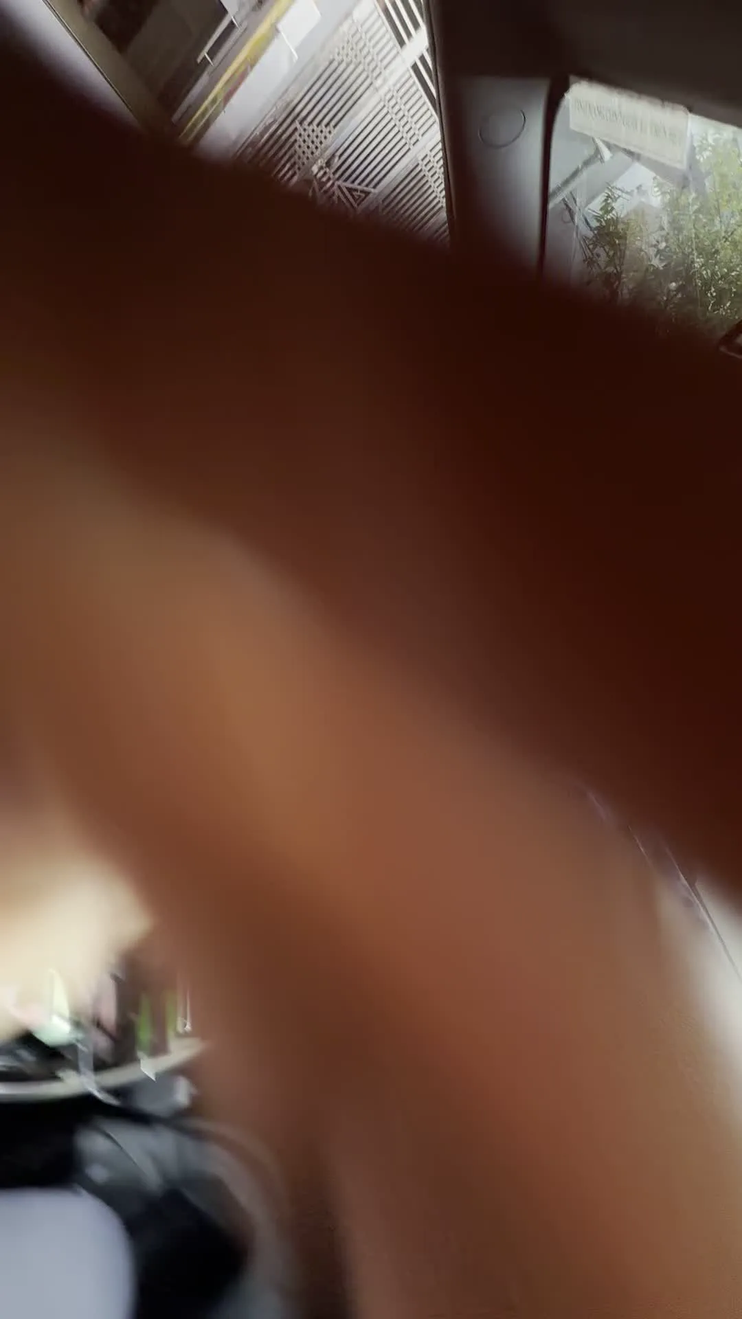 Quang cảnh được ghi lại từ bên trong một chiếc ô tô hiện đại. Một người đàn ông đeo kính, mặc áo khoác ghi lê, đang ngồi ở vị trí lái xe và cầm vô lăng. Nội thất của xe có thể được nhìn thấy rõ, bao gồm vô lăng có logo hình chữ 'V' (dường như là xe VinFast), một màn hình cảm ứng lớn đặt dọc ở bảng điều khiển trung tâm, và một chiếc điện thoại thông minh được gắn trên táp-lô. Nhìn qua kính chắn gió và cửa sổ bên, có thể thấy khung cảnh đường phố với các tòa nhà và một phương tiện khác. Có chữ viết trên gương chiếu hậu nhưng bị ngược và không thể đọc được.