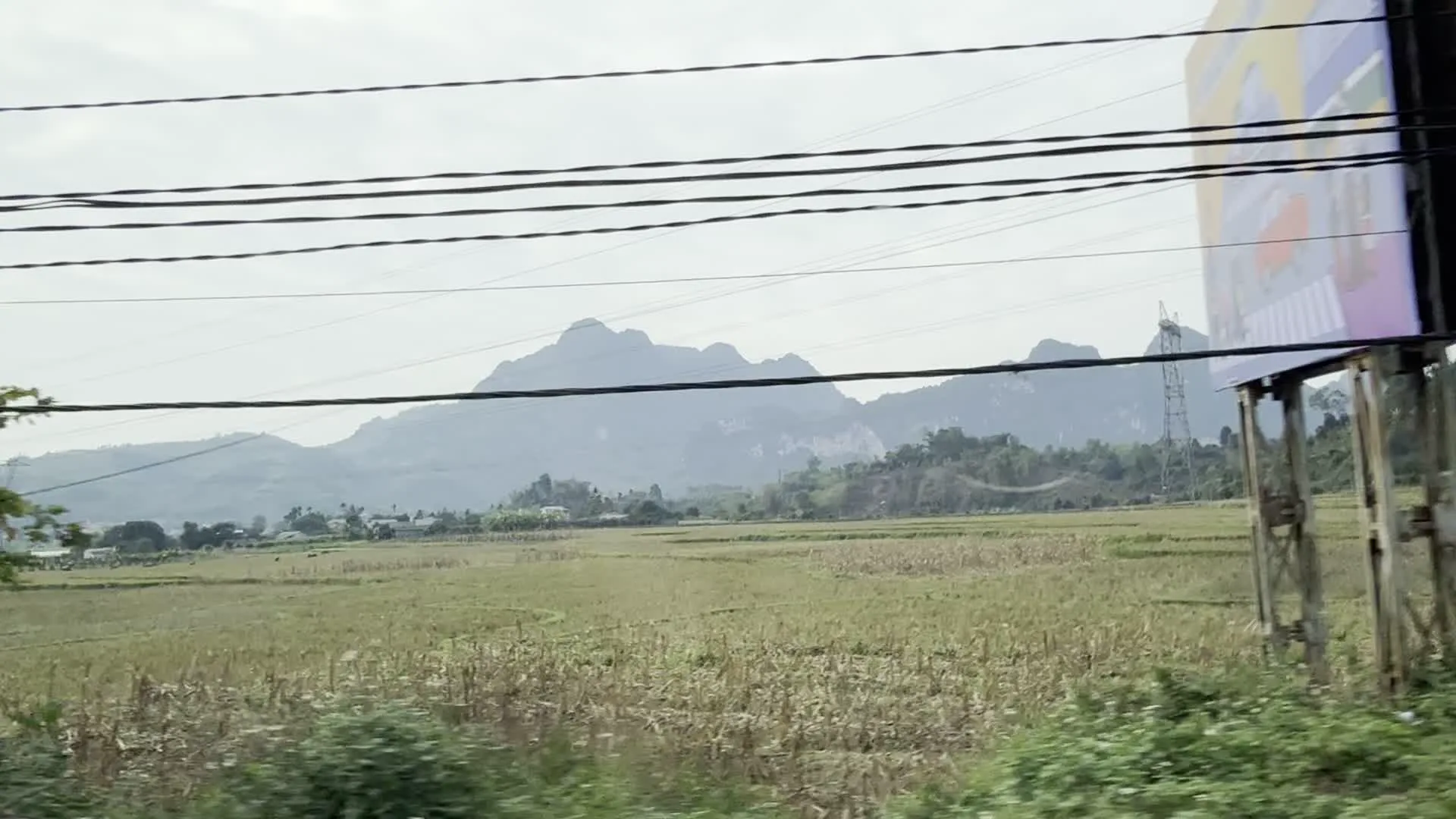 Cảnh vật được ghi lại từ một phương tiện đang di chuyển, cho thấy phong cảnh nông thôn đặc trưng ở vùng trung du miền núi, có thể là tại tỉnh Hòa Bình. Hình ảnh nổi bật là một cánh đồng rộng lớn, có lẽ là ruộng lúa đã gặt xong, trải dài đến chân một dãy núi đá vôi hùng vĩ ở phía xa. Bầu trời mờ ảo và có nhiều mây. Nhiều đường dây điện giăng ngang qua khung hình ở phía trên. Dọc theo con đường là những ngôi nhà dân. Một số ngôi nhà có kiến trúc hiện đại với cổng và hàng rào sắt, trong khi những ngôi nhà khác mang dáng vẻ nhà cấp bốn truyền thống ở nông thôn. Bên lề đường có thể thấy cây cối và một vài biển quảng cáo, tuy nhiên nội dung trên các biển này không thể đọc được do ảnh chụp bị mờ và rung lắc khi di chuyển. Không có hoạt động cụ thể nào của con người được ghi lại, chủ yếu là quang cảnh tĩnh lặng của làng quê.