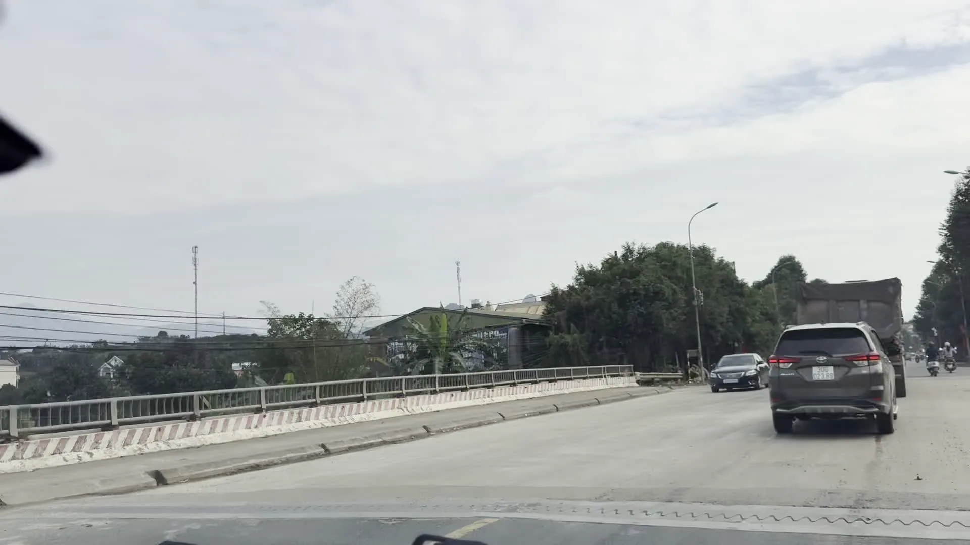 Khung cảnh được ghi lại từ một chiếc ô tô đang di chuyển trên một cây cầu ở khu vực có dân cư sinh sống, có thể thuộc tỉnh Hòa Bình. Trên đường có nhiều phương tiện đang lưu thông, bao gồm ô tô, xe tải và xe máy. Hai bên đường là cây cối, nhà dân, cột đèn và có một lan can cầu bằng bê tông với vạch sơn màu đỏ và trắng. Phía xa là những ngọn đồi. Bên phải đường có một tấm biển lớn tuyên truyền về an toàn giao thông với dòng chữ "AN TOÀN GIAO THÔNG LÀ HẠNH PHÚC CỦA..." cùng hình ảnh minh họa một gia đình đang qua đường.