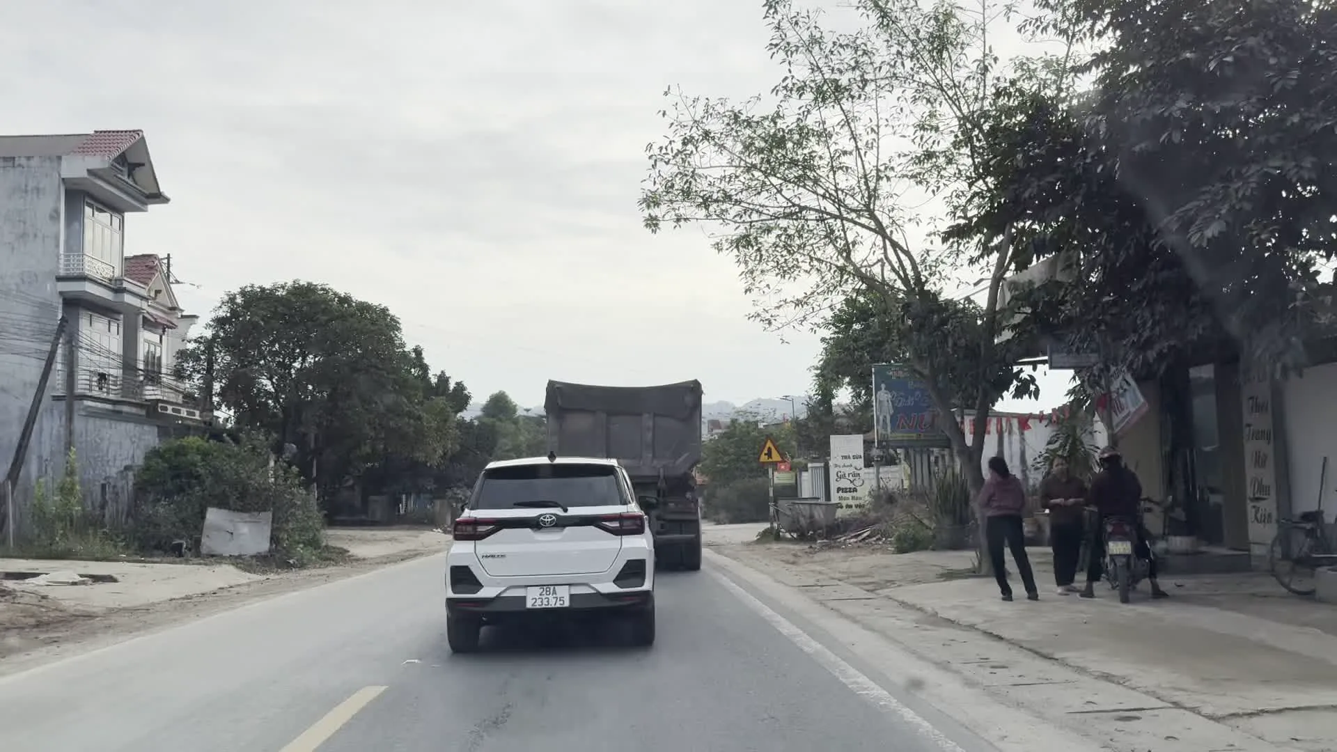 Cảnh quay từ bên trong một chiếc ô tô đang di chuyển trên một con đường ở khu vực nông thôn hoặc ngoại ô. Một chiếc ô tô SUV màu trắng đang đi phía trước, cùng với các phương tiện khác như xe tải và xe máy. Hai bên đường là nhà dân và các cửa hàng. Có thể đọc được một số biển hiệu như một tiệm làm tóc có chữ 'TÓC', và một xưởng cơ khí có biển hiệu 'CƠ KHÍ HIẾU LAN' và 'NHÔM KÍNH - CỬA KÍNH - CỬA SẮT'. Bên lề đường, có một nhóm người đang đứng và một vài chiếc xe máy đang đậu. Một ngôi nhà treo cờ Việt Nam. Phía xa có thể thấy những ngọn núi dưới bầu trời u ám.