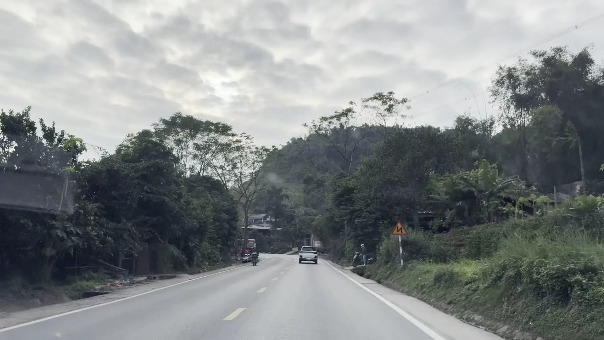 Đây là khung cảnh trên một con đường nhựa ở khu vực miền núi, có thể là ở tỉnh Hòa Bình. Bầu trời u ám và nhiều mây. Con đường có hai làn xe, uốn lượn qua những ngọn đồi được bao phủ bởi cây cối rậm rạp, bao gồm tre, nứa và cây chuối. Một số phương tiện đang lưu thông trên đường, bao gồm một chiếc ô tô bán tải màu trắng, một xe tải, một xe khách nhỏ màu trắng và một chiếc xe ba gác đang di chuyển ở lề đường. Có các biển báo giao thông ven đường, bao gồm một biển báo nguy hiểm hình tam giác màu vàng cảnh báo đường cong và một biển báo giới hạn tốc độ 40 km/h. Ở một đoạn, có một chiếc ô lớn có thể là của một quán ven đường và một tấm biển màu xanh có chữ trắng không thể đọc rõ. Hệ thống cột điện và đường dây điện cao thế cũng xuất hiện dọc theo tuyến đường và trên sườn đồi.