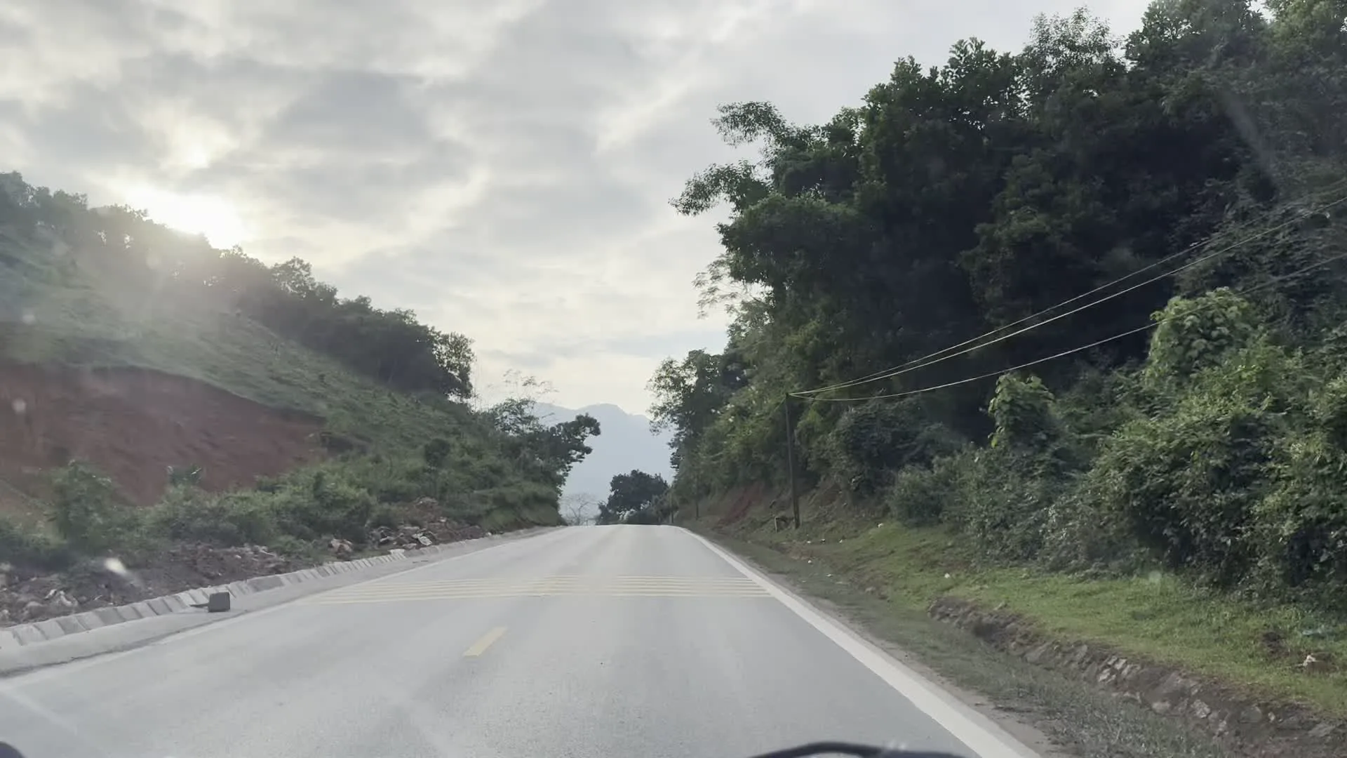 Đây là những hình ảnh ghi lại khung cảnh trên một con đường đèo ở khu vực miền núi tại Hòa Bình. Con đường được trải nhựa, có vạch kẻ đường màu vàng ở giữa, uốn lượn qua những ngọn đồi. Hai bên đường là cảnh quan thiên nhiên với cây cối um tùm, bao gồm cả những lùm tre, nứa. Một bên là sườn đồi dốc với thảm thực vật xanh tươi, có nơi là vách đất màu nâu đỏ. Phía xa là những dãy núi trập trùng ẩn hiện dưới bầu trời nhiều mây. Mặt trời đôi lúc ló dạng qua các đám mây, chiếu sáng một vùng trời. Ven đường có một vài ngôi nhà dân trông đơn sơ với mái lợp tôn và có cả đường dây điện chạy dọc theo. Trong một bức ảnh, có thể thấy một biển báo giao thông ở phía xa nhưng không thể đọc rõ nội dung. Toàn bộ các hình ảnh đều được chụp từ góc nhìn của một người đang ngồi trong xe ô tô di chuyển trên đường.