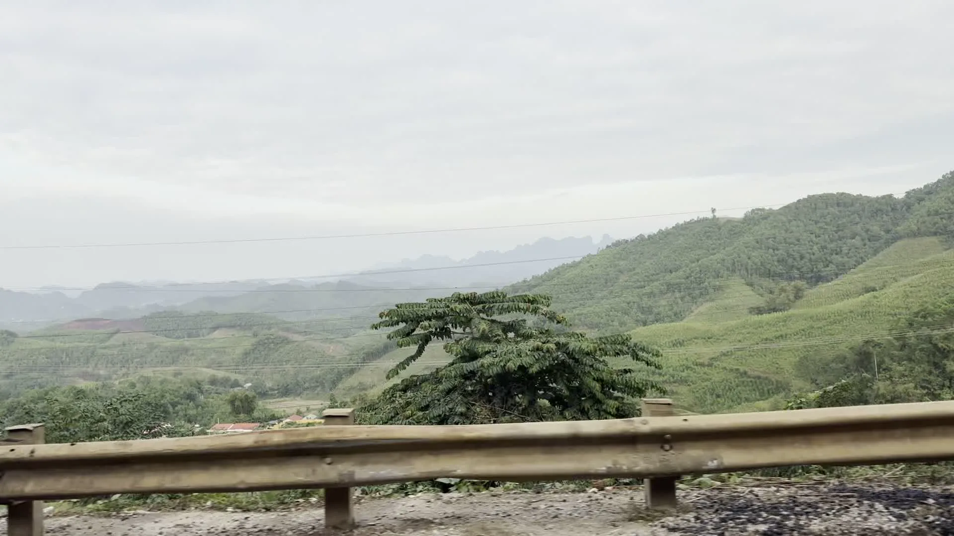 Bức ảnh chụp phong cảnh một vùng đồi núi, được nhìn từ một con đường. Ở phía trước, có một hàng rào hộ lan bằng kim loại chạy dọc theo con đường. Phía sau hàng rào là một khung cảnh rộng lớn với những ngọn đồi và dãy núi trập trùng, được bao phủ bởi cây cối xanh tươi. Có thể thấy các đường dây điện chạy ngang qua sườn đồi. Bầu trời có màu xám và nhiều mây, cho thấy thời tiết đang u ám. Ở phía thung lũng xa xa, có thể thấp thoáng thấy một vài mái nhà. Không có hoạt động cụ thể nào của con người được ghi lại, chỉ có cảnh quan thiên nhiên và một phần cơ sở hạ tầng giao thông.