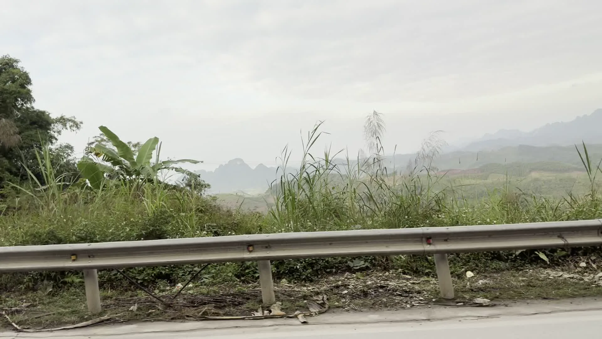Đây là khung cảnh được chụp từ một chiếc xe đang di chuyển trên một con đường ở vùng nông thôn, miền núi. Dọc theo con đường là lan can bảo vệ bằng kim loại và bê tông. Bên vệ đường có cây cỏ mọc um tùm, trong đó có nhiều cây chuối. Một bức tường xây bằng gạch bê tông cũng xuất hiện. Phía xa là những dãy núi trập trùng mờ ảo trong sương mù dưới bầu trời u ám. Có thể thấy dây điện chạy ngang qua. Thấp thoáng sau lùm cây có một tấm bạt màu xanh. Không có hoạt động nào của con người được ghi lại và không có văn bản hay chữ viết nào.