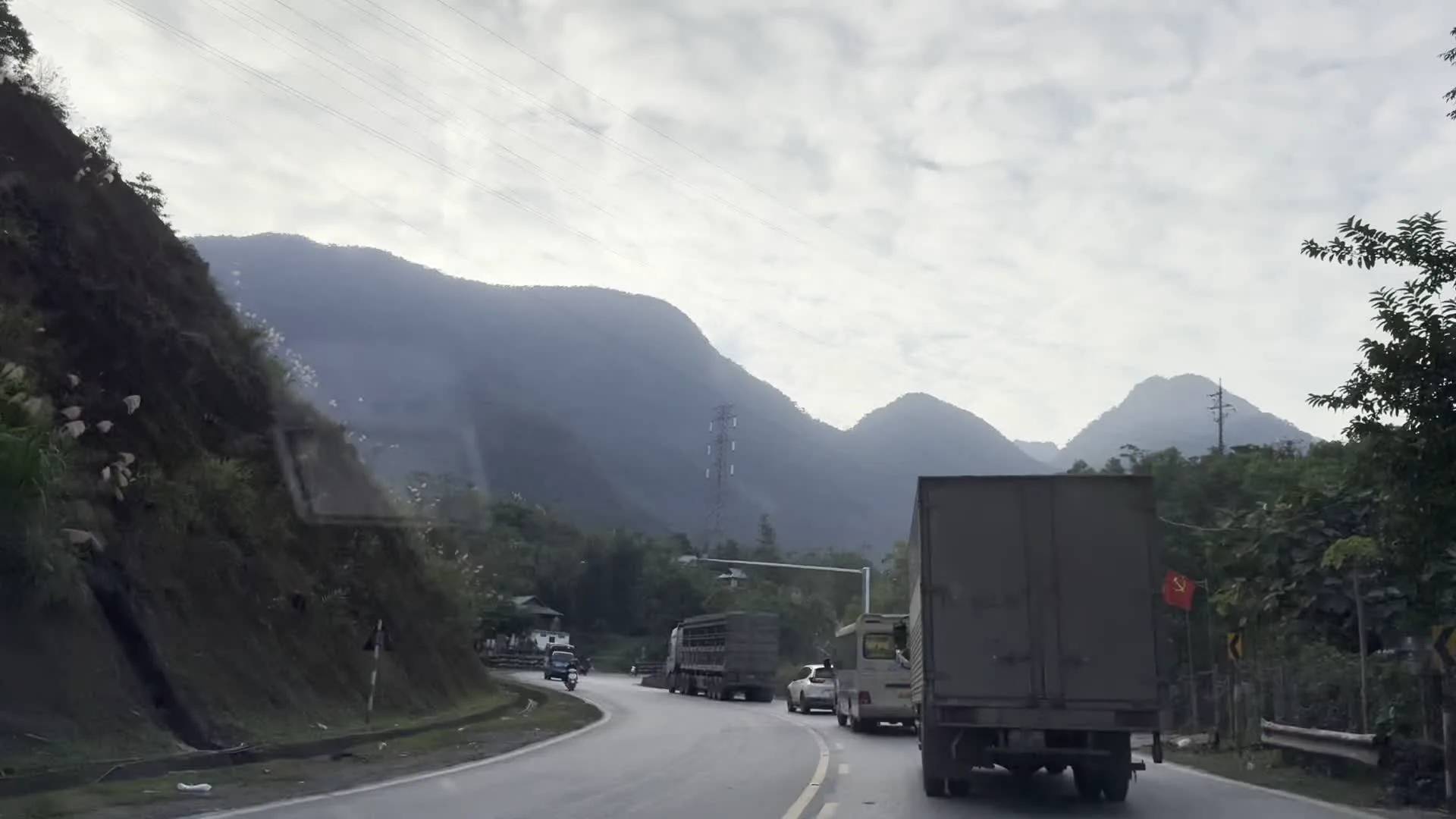 Khung cảnh được ghi lại từ một phương tiện đang di chuyển trên một con đường nhựa quanh co ở vùng núi. Dọc đường có nhiều phương tiện giao thông như xe tải, xe ô tô con, xe khách nhỏ và xe máy. Đi ngay phía trước là một chiếc xe tải lớn màu xanh, phía sau có cắm lá cờ đỏ sao vàng. Phong cảnh hai bên đường chủ yếu là đồi núi trập trùng, cây cối xanh tươi và sương mù bao phủ phía xa. Bầu trời âm u, nhiều mây. Có thể nhìn thấy hệ thống đường dây điện và một cột điện cao thế. Ven đường có một vài ngôi nhà của người dân, trong đó có một ngôi nhà sàn. Một biển báo giao thông hình tam giác và một tấm biển hiệu có chữ 'OTO' cũng xuất hiện nhưng không thể đọc rõ toàn bộ nội dung.