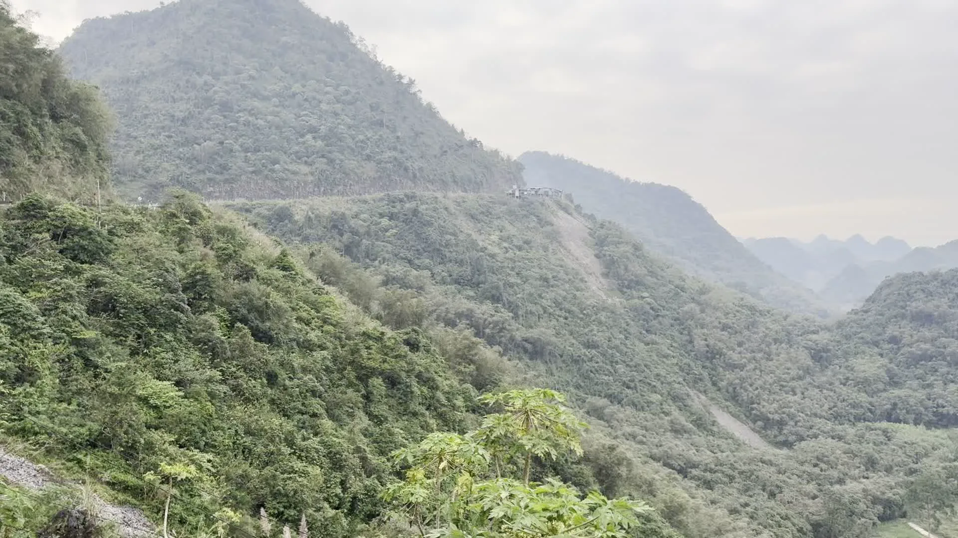 Quang cảnh được chụp tại một vùng núi ở Hòa Bình, đặc trưng bởi những dãy núi đá vôi trùng điệp được cây cối xanh tươi bao phủ. Bầu trời có nhiều mây và không khí có vẻ hơi mờ sương.。Trong ảnh, có một con đường đèo quanh co chạy dọc theo sườn núi. Nhìn từ trên cao xuống thung lũng là các khu dân cư thưa thớt với những ngôi nhà nằm rải rác, xen kẽ là các thửa ruộng và đường đất. Một cột điện cao thế có thể được nhìn thấy trên một đỉnh đồi.。Đáng chú ý là một cụm các công trình và nhà cửa được xây dựng trên một sườn núi dốc đã được san gạt, cho thấy có thể là một khu định cư nhỏ, một công trường khai thác hoặc một điểm dừng chân trên núi. Một đường dây cáp điện vắt ngang qua một trong các bức ảnh. Toàn cảnh thể hiện một khung cảnh thiên nhiên hùng vĩ, yên bình của vùng nông thôn miền núi Việt Nam. Không có bất kỳ chữ viết nào có thể đọc được trong các hình ảnh.