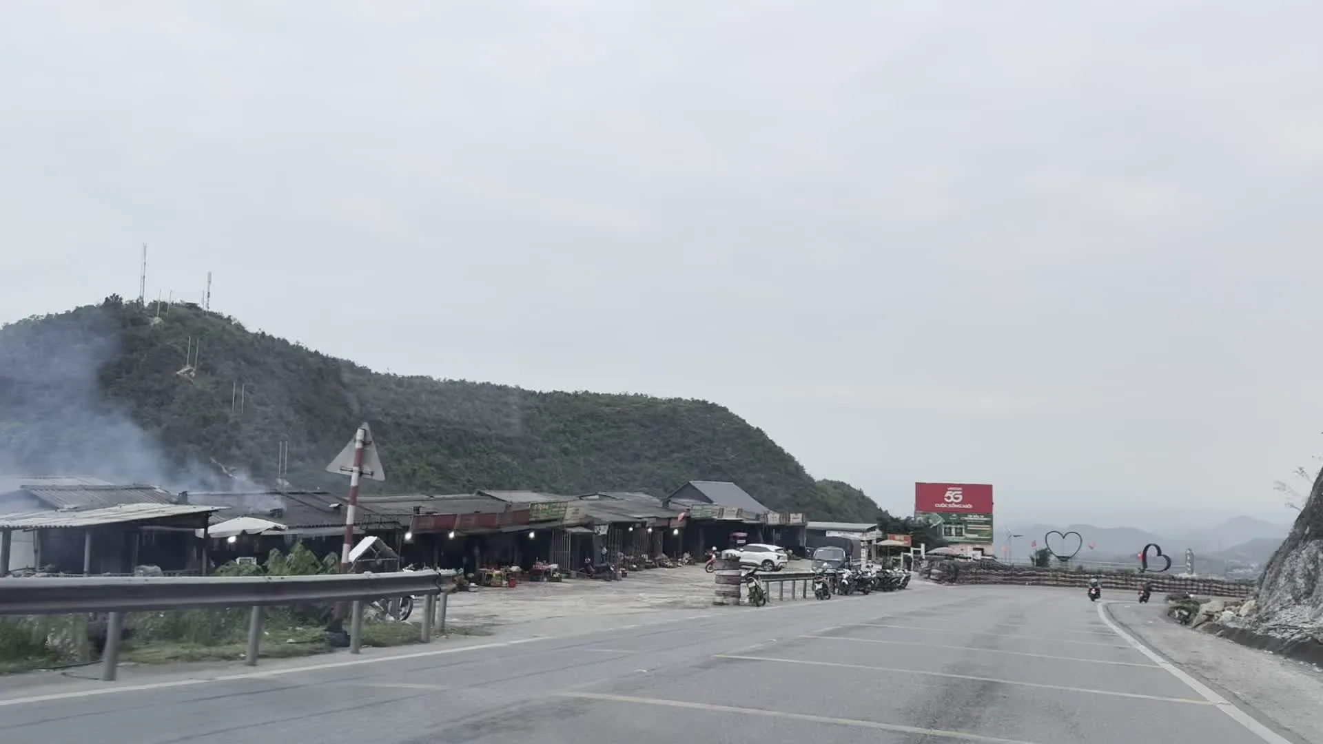 Cảnh quan ven một con đường đèo ở vùng núi. Có một dãy các quán ven đường lợp mái tôn, trông giống như một khu chợ nhỏ hoặc trạm dừng chân. Phía trước các quán có nhiều xe máy và một chiếc ô tô màu trắng đang đỗ. Có khói bốc lên từ một trong các ngôi nhà, có thể là khói nấu ăn. Ở phía xa bên phải, có một biển quảng cáo lớn màu đỏ có chữ '5G' và một biểu tượng trang trí hình trái tim. Con đường được bảo vệ bởi hàng rào làm từ các lốp xe cũ xếp chồng lên nhau. Con đường uốn lượn quanh sườn núi, với một bên là vách núi đá và một bên là sườn núi được bao phủ bởi cây cối rậm rạp.