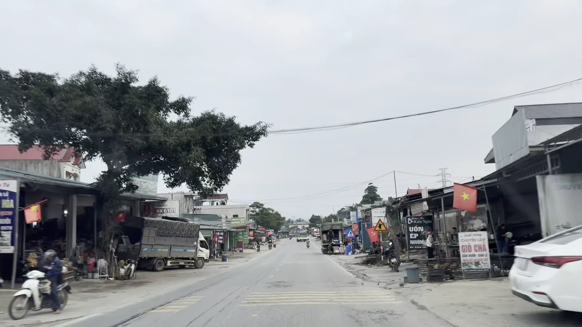 Khung cảnh dọc một con đường ở khu vực nông thôn hoặc thị trấn của Việt Nam. Hai bên đường là những ngôi nhà và cửa hàng san sát nhau. Có nhiều phương tiện giao thông như xe máy, ô tô và xe tải đang di chuyển và đậu ven đường. Nhiều ngôi nhà treo cờ Việt Nam. Có thể đọc được một số biển hiệu của các cửa hàng, chẳng hạn như một quán bán 'GIÒ CHẢ', một tiệm làm tóc có chữ 'UỐN ÉP' và một biển hiệu khác ghi 'Kiên Ngân'. Trên một biển hiệu khác có chữ 'MUA THU'. Cảnh vật thay đổi từ khu dân cư có hàng quán sang một đoạn đường thoáng đãng hơn với cây cối và đồi núi ở hai bên.