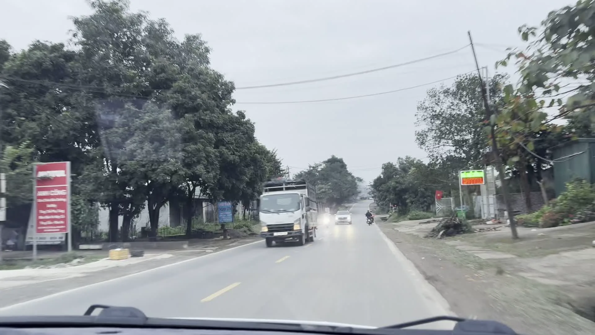 Quang cảnh được ghi lại từ góc nhìn bên trong một chiếc ô tô đang di chuyển trên một con đường nhựa ở khu vực có vẻ là nông thôn hoặc ngoại ô. Bầu trời âm u. Hai bên đường có nhiều cây xanh. Có nhiều phương tiện giao thông đang di chuyển trên đường, bao gồm một chiếc xe tải màu trắng đang bật đèn pha, xe ô tô con, và một vài người đang đi xe máy cùng một chiếc xe ba gác. Ven đường có một số tòa nhà và cửa hàng. Có một tấm biển quảng cáo màu đỏ ở bên đường nhưng chữ viết không thể đọc rõ. Trong một hình ảnh, có thể thấy một lá cờ Việt Nam được treo ở cổng một ngôi nhà.