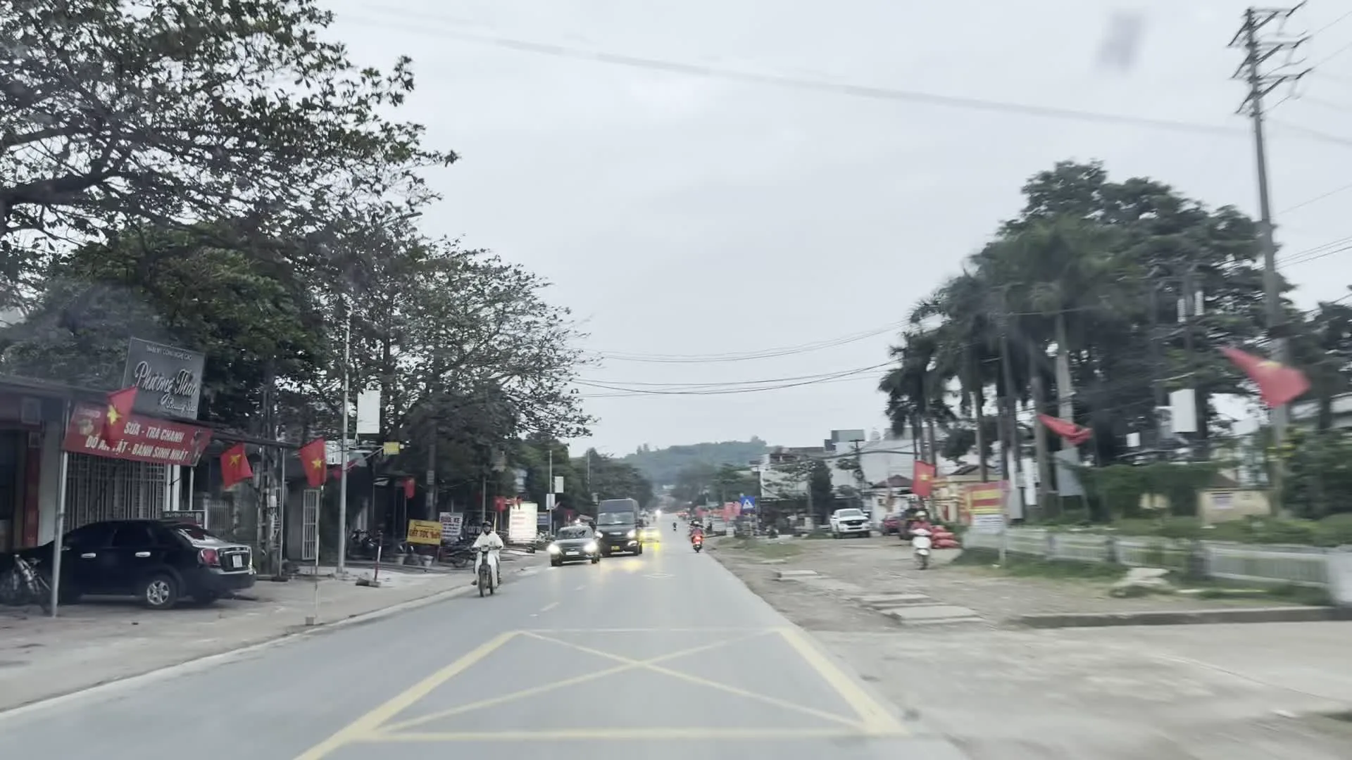 Quang cảnh một con đường tại một thị trấn hoặc khu vực nông thôn ở Việt Nam, nhìn từ trong một chiếc ô tô. Con đường có nhiều phương tiện giao thông qua lại, bao gồm ô tô, xe tải và xe máy. Hai bên đường là các ngôi nhà, cửa hàng san sát nhau. Nhiều lá cờ đỏ sao vàng của Việt Nam được treo trước các ngôi nhà và cửa hàng. Có một số biển hiệu, một vài trong số đó có thể đọc được như "Phượng Sài Gòn", "Sơn - Sửa Chữa ... ĐIỆN - LẠNH - DÂN DỤNG" và một biển hiệu khác có chữ "RỬA XE". Cảnh vật cho thấy hoạt động sinh hoạt và kinh doanh thường ngày. Bầu trời có mây, u ám.