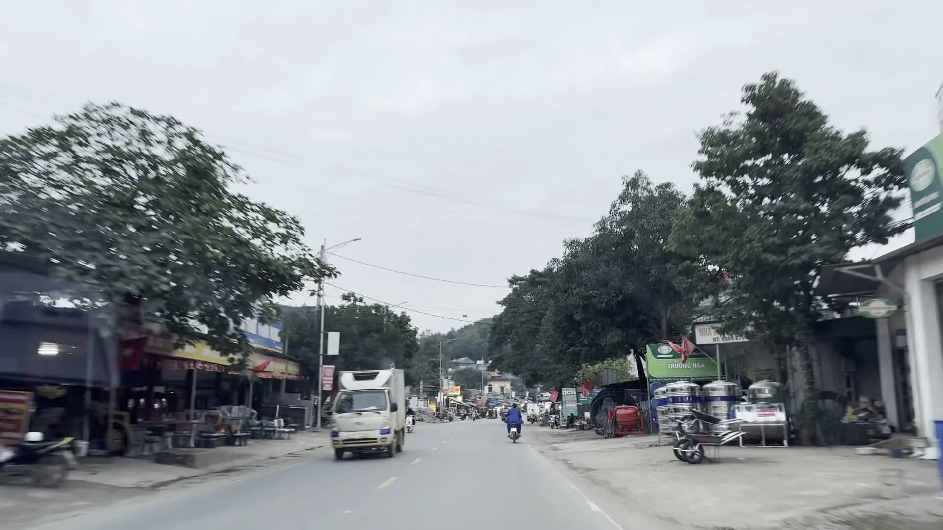 Quang cảnh một con đường ở khu vực thị trấn hoặc nông thôn tại Việt Nam, có thể ở Hòa Bình, dưới bầu trời nhiều mây. Hai bên đường là các cửa hàng, nhà dân với nhiều biển hiệu quảng cáo. Một số biển hiệu có thể đọc được chữ tiếng Việt như "VÀNG BẠC", "BIỂN QUẢNG CÁO", "HOA HỒNG". Trên đường có các phương tiện giao thông đang di chuyển, bao gồm xe tải nhỏ, xe máy và ô tô con. Hoạt động của người dân diễn ra dọc hai bên đường; có hình ảnh trông giống một khu chợ nhỏ hoặc các hàng quán ven đường, nơi mọi người đang tụ tập. Có nhiều cây xanh lớn dọc hai bên đường và cờ Việt Nam được treo ở một số nơi.