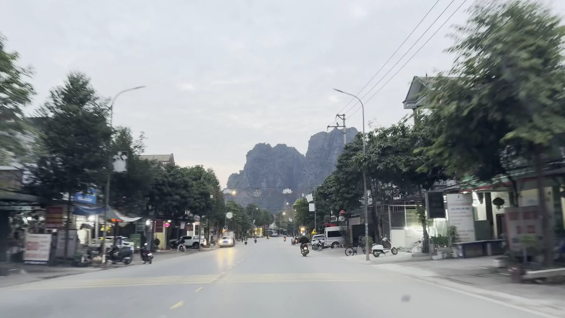 Quang cảnh được ghi lại từ một chiếc xe đang di chuyển trên một con đường ở một thị trấn miền núi, có nền là những dãy núi đá vôi hùng vĩ. Bầu trời nhiều mây, có thể là vào buổi sáng sớm hoặc chiều tà. Hai bên đường là nhà dân, các cửa hàng và cây xanh. Có một số phương tiện giao thông như xe máy, ô tô và xe tải đang lưu thông. Trên đường có các công trình trang trí, bao gồm một cổng chào lớn với họa tiết rồng mây và một biểu tượng hoa sen phát sáng ở phía xa. Có thể nhìn thấy một số biển hiệu và biểu ngữ, nhưng chữ viết không thể đọc rõ, ngoại trừ một tấm biển có chữ "BÚN CHẢ".