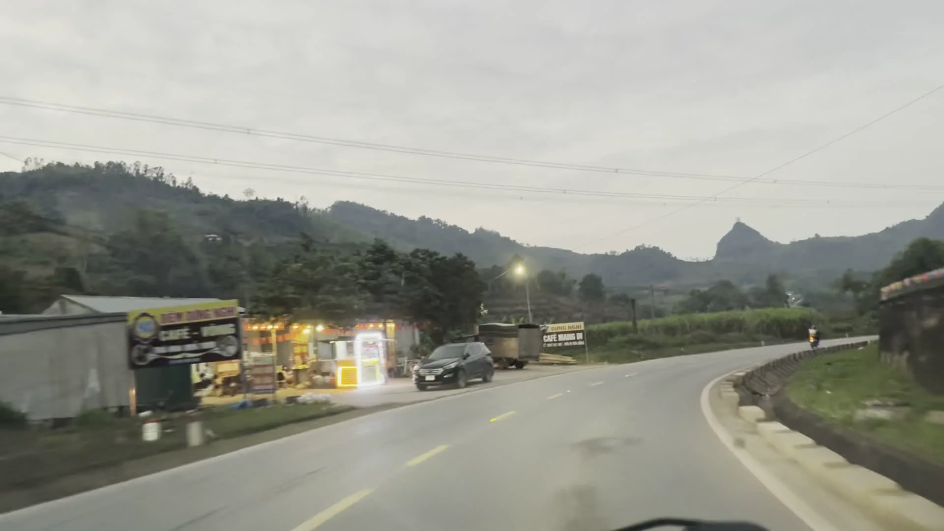 Cảnh quay từ bên trong một chiếc ô tô đang di chuyển trên một con đường quốc lộ ở khu vực miền núi, có thể là vào lúc chạng vạng hoặc sáng sớm. Khung cảnh cho thấy nhiều phương tiện giao thông và các hoạt động ven đường.。Các thông tin từ biển hiệu có thể đọc được:。- Một biển hiệu lớn màu vàng ghi "ĐIỂM DỪNG NGHỈ" và bên dưới là "CAFE - Lẩu".。- Một biển hiệu khác gần đó ghi "ĐƯỜNG NGANG" và "CAFE MẠNH VI".。- Một biển hiệu màu vàng khác ghi "RỬA XE".。Hoạt động và cảnh vật quan sát được:。- Địa điểm: Một con đường nhựa ở vùng nông thôn, miền núi với đồi núi trập trùng ở phía xa. Có một công trình kiến trúc hoặc tượng đài trên đỉnh một ngọn núi.。- Hoạt động: Nhiều phương tiện đang tham gia giao thông, bao gồm ô tô con, xe máy, và một chiếc xe tải bồn lớn (có chữ SHACMAN ở đầu xe). Các phương tiện đều bật đèn pha. Các quán ăn, quán cà phê và tiệm rửa xe ven đường đang mở cửa hoạt động.。- Vật thể: Các ngôi nhà cấp 4 và quán xá ven đường, một chiếc ô tô màu đen đang đỗ, cây cối rậm rạp, cột điện, và một lá cờ Việt Nam được treo trước một ngôi nhà.