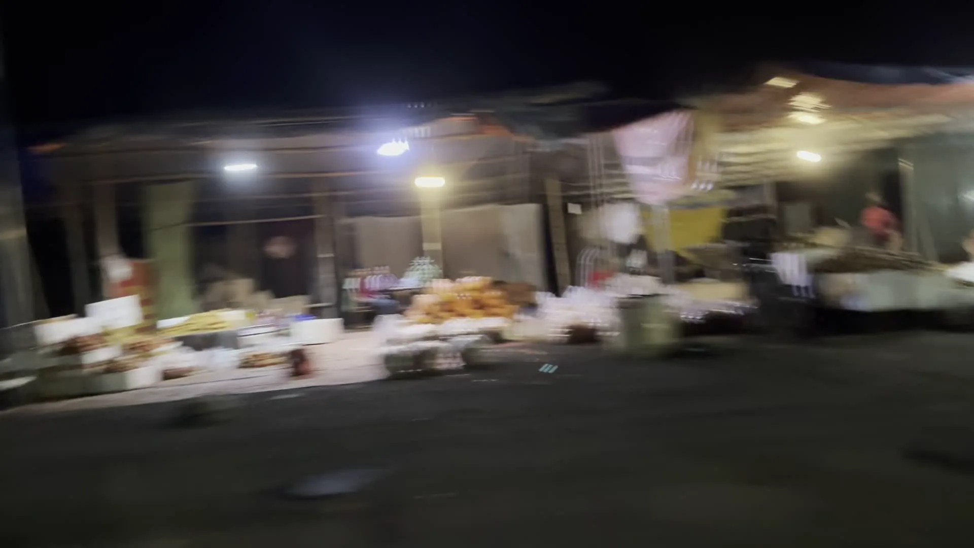 Hình ảnh bị mờ về một khu chợ ven đường vào ban đêm, được chụp từ một phương tiện đang di chuyển.