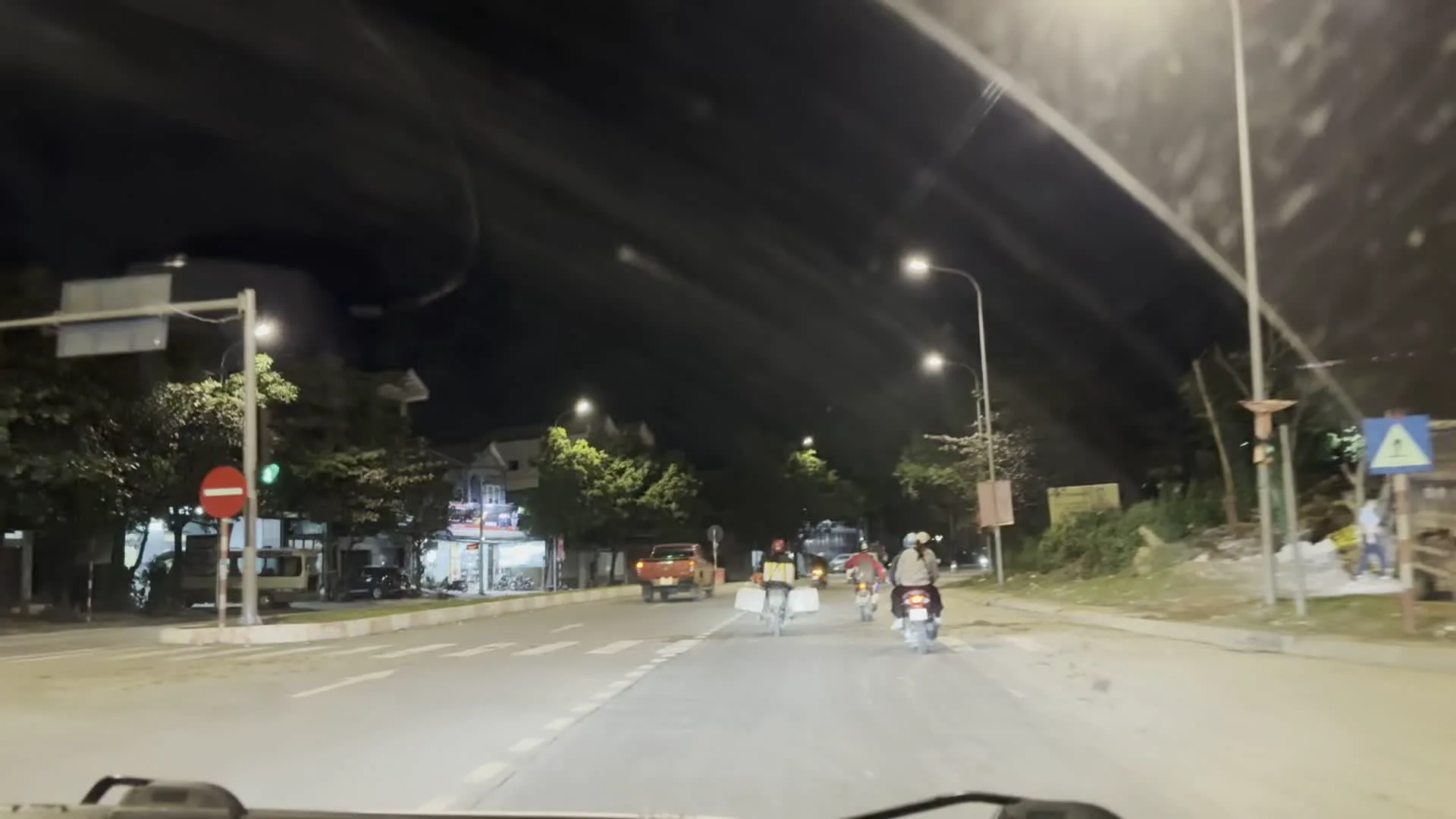 Cảnh quay vào ban đêm trên một con đường nhựa trong đô thị, nhìn từ góc nhìn của người lái xe ô tô. Con đường được chiếu sáng bởi hệ thống đèn đường. Có nhiều phương tiện đang lưu thông, bao gồm ô tô, xe máy và một người đi xe đạp chở thùng hàng phía sau, có khả năng là đang đi giao hàng. Hai bên đường có cây xanh và các tòa nhà, cửa hàng san sát. Một số biển hiệu quảng cáo được dựng lên nhưng không thể đọc rõ chữ. Trong một khung hình có thể nhìn thấy một cột đèn tín hiệu giao thông đang bật đèn đỏ và một biển báo giao thông.