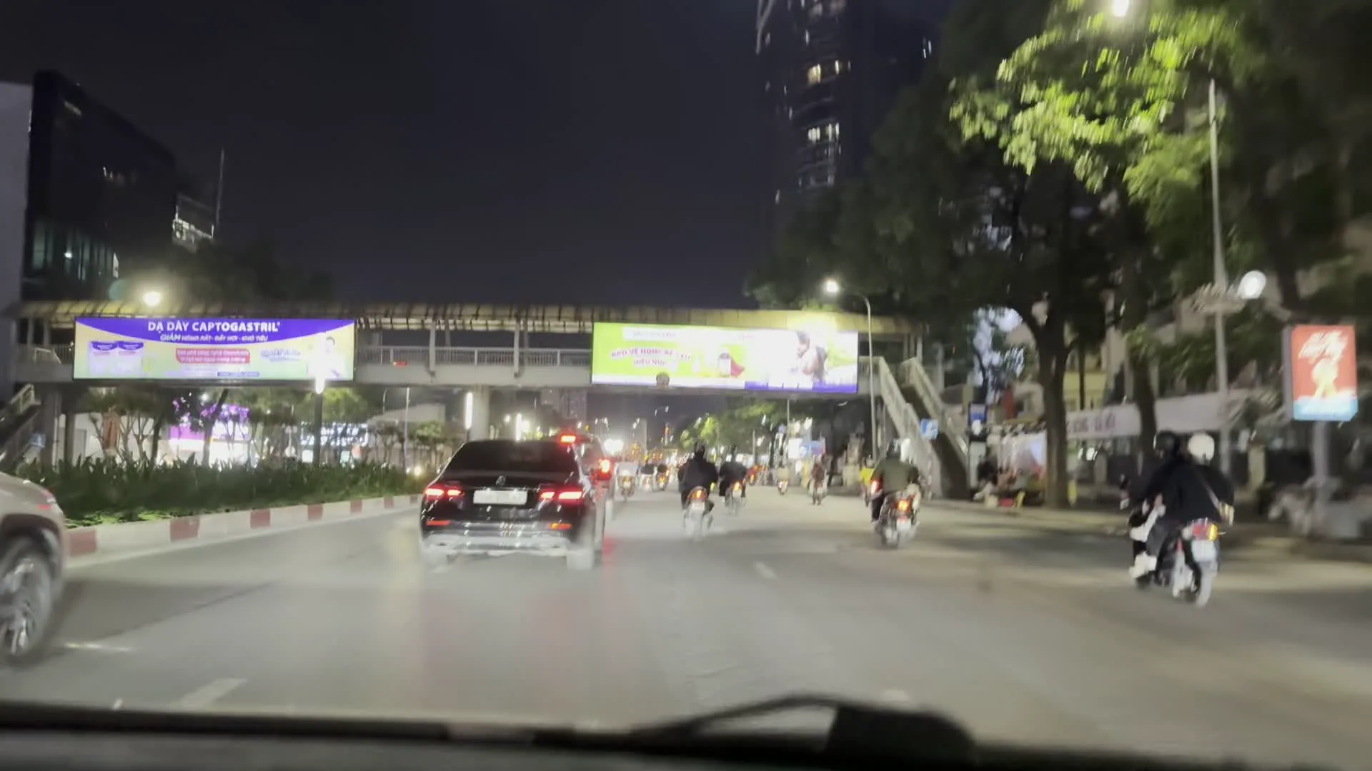 Quang cảnh vào ban đêm trên một con đường trong thành phố, nhìn từ bên trong một chiếc ô tô đang di chuyển. Trên đường có nhiều phương tiện giao thông như ô tô và xe máy. Hai bên đường là các tòa nhà cao tầng hiện đại và cây xanh. Có một cây cầu vượt dành cho người đi bộ, trên đó có gắn các biển quảng cáo lớn, trong đó một biển có thể đọc được chữ "DẠ DÀY CAPTOGASTRIL". Ngoài ra, một tòa nhà cao tầng có biển hiệu nổi bật với chữ "D.CHIC". Đường phố rộng, có nhiều làn xe và được chiếu sáng bởi đèn đường.