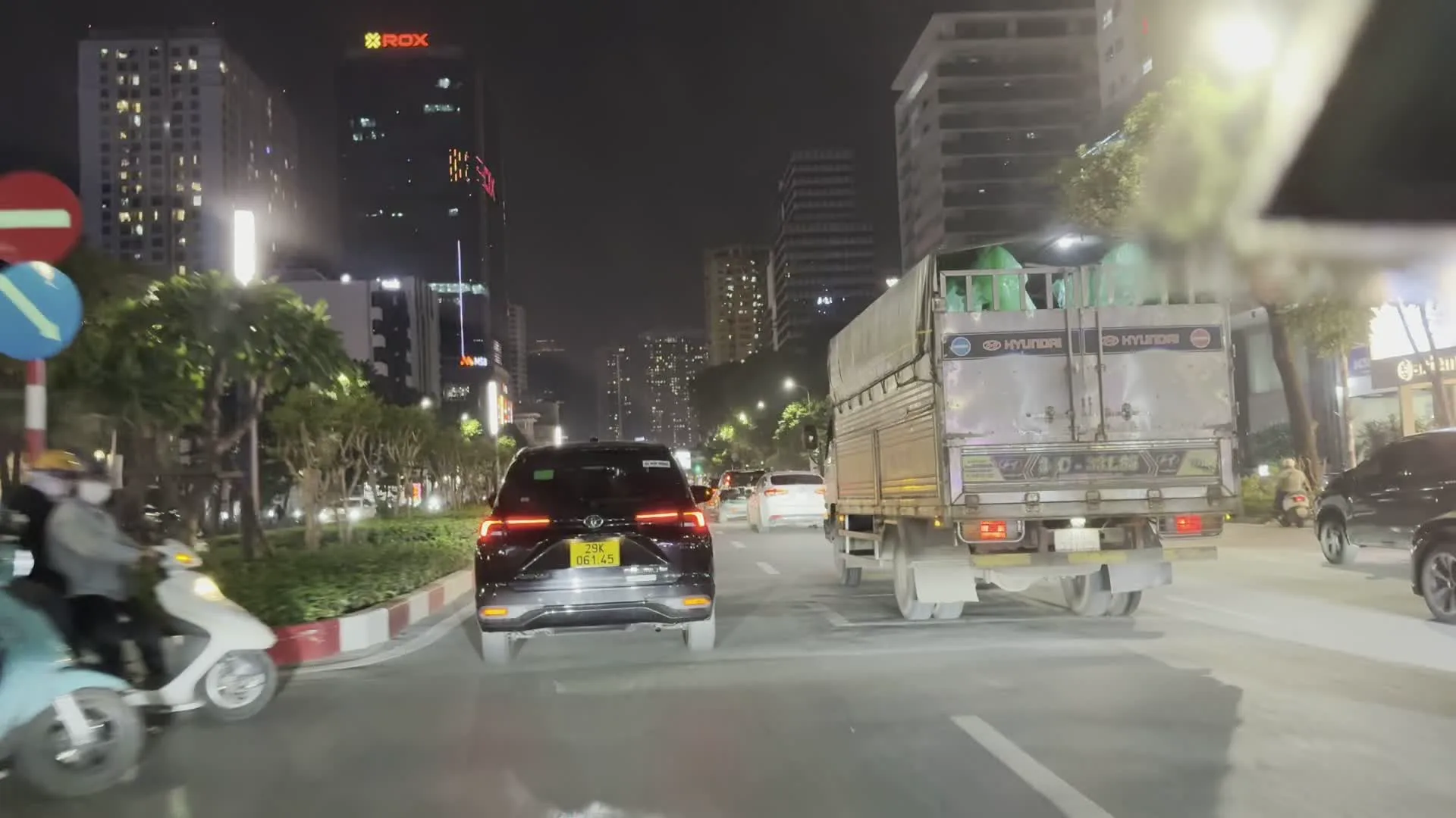 Quang cảnh một con đường đô thị vào ban đêm với nhiều phương tiện giao thông đang lưu thông, bao gồm ô tô, xe máy và một chiếc xe tải. Hai bên đường là các tòa nhà cao tầng hiện đại được thắp sáng. Có một biển báo cấm đi ngược chiều và một biển báo hình tròn màu xanh. Có các biển quảng cáo và biển hiệu, trong đó có một biển hiệu trên tòa nhà ghi 'ROX', và một biển quảng cáo lớn bắc qua đường với các dòng chữ 'MSB' và 'CĂN HỘ NGHỈ DƯỠNG'.