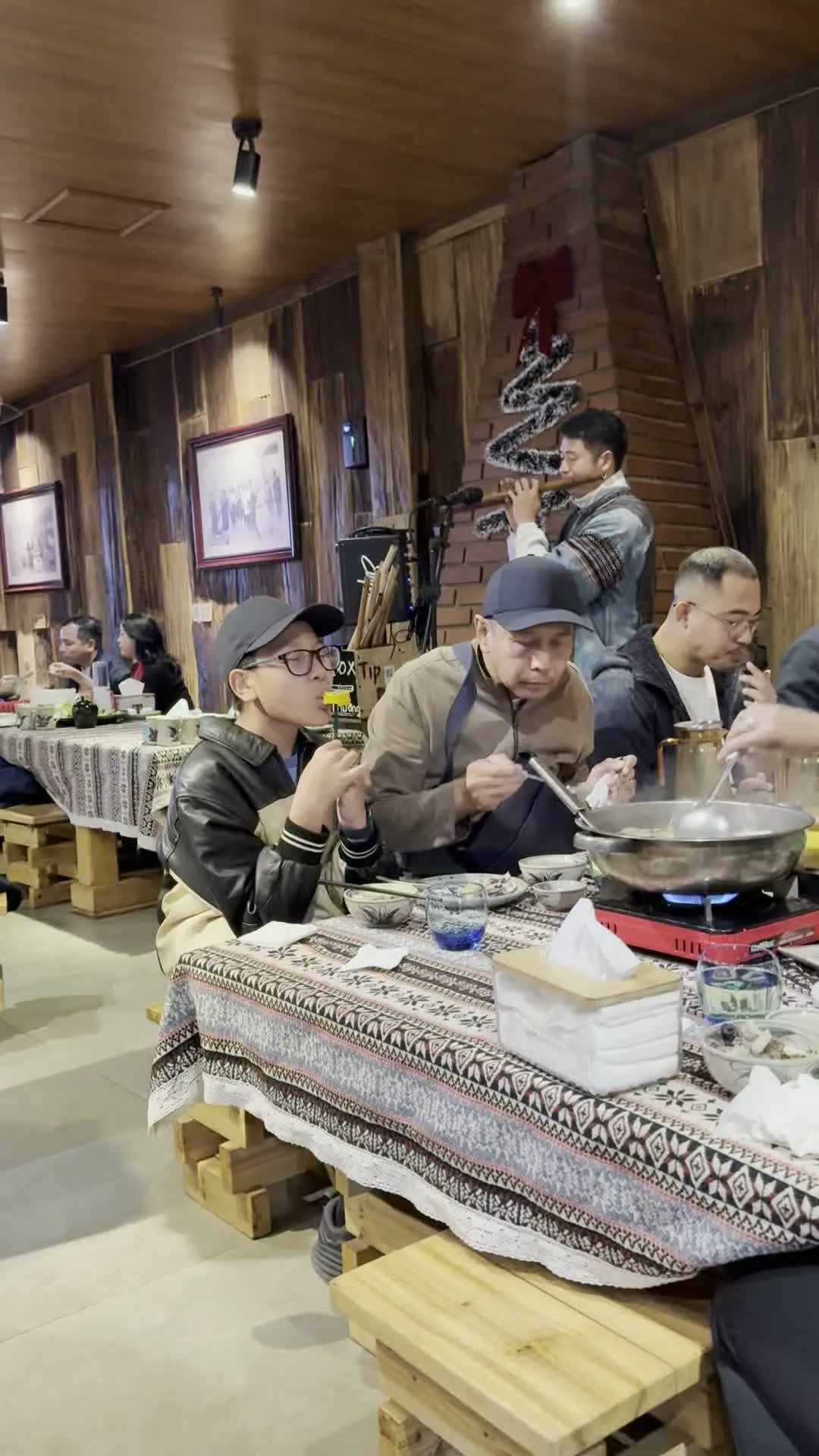 Bối cảnh là bên trong một nhà hàng với nội thất và tường chủ yếu bằng gỗ, tạo cảm giác mộc mạc, ấm cúng. Một nhóm người đang quây quần bên bàn ăn và thưởng thức lẩu. Trên bàn, một nồi lẩu lớn đang sôi được đặt trên bếp ga mini. Mọi người đang dùng bát và đũa để ăn. Bàn được trải một tấm khăn có hoa văn thổ cẩm đặc trưng. Trong số các thực khách có một cậu bé đeo kính, đội mũ lưỡi trai, và những người đàn ông khác. Ở phía sau, một nhạc công đang đứng thổi sáo, biểu diễn cho các thực khách. Trên tường có treo những bức ảnh đen trắng được đóng khung và có một chiếc hộp ghi chữ "BOX TIP".