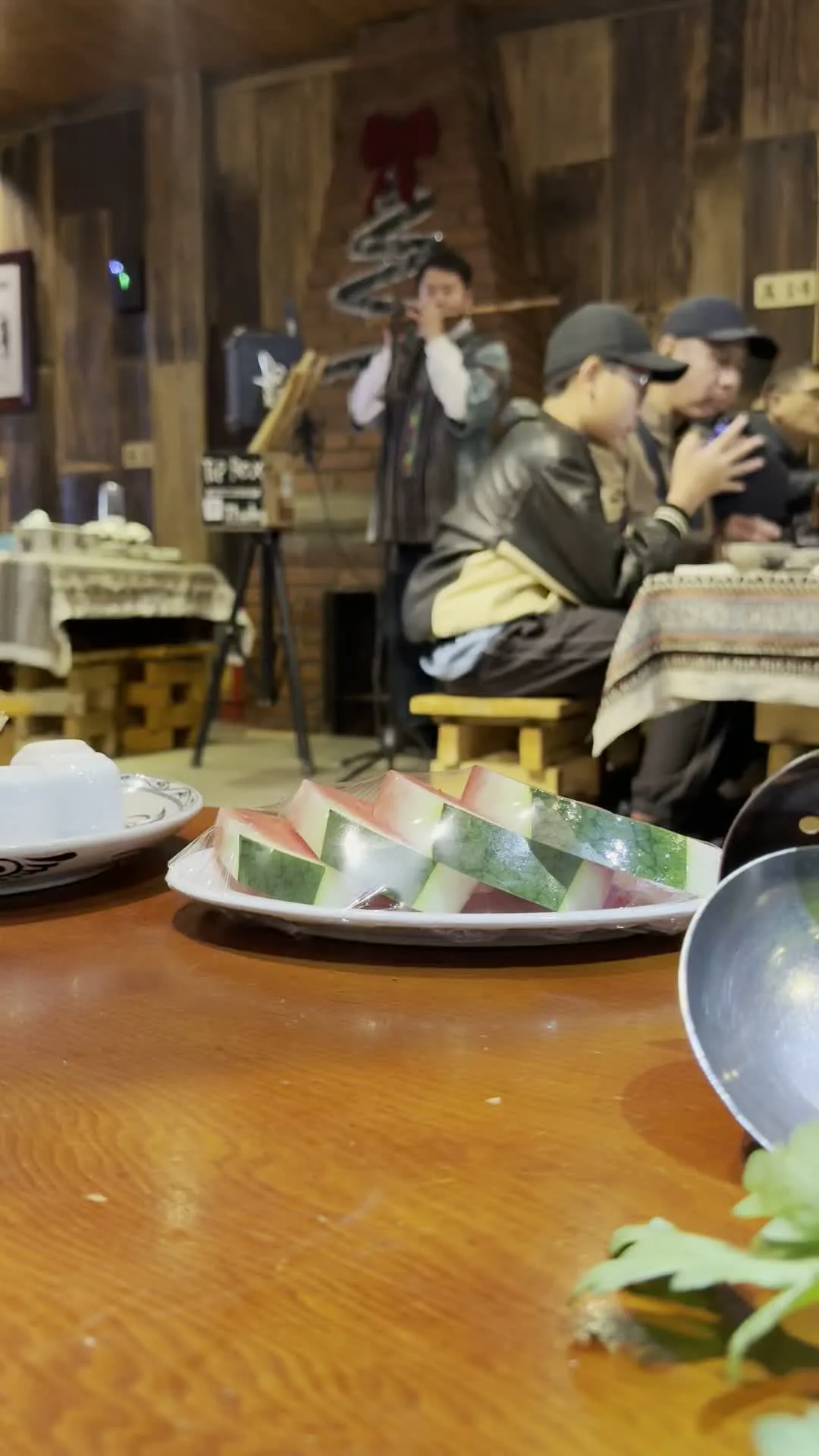 Khung cảnh bên trong một nhà hàng có tường và đồ nội thất bằng gỗ, tạo cảm giác mộc mạc. Ở phía sau, một nhạc công đang đứng biểu diễn một loại nhạc cụ hơi, có thể là sáo hoặc khèn, cho thực khách xem. Trước mặt nhạc công có một micrô trên giá đỡ. Một nhóm khách đang ngồi ăn tại bàn và thưởng thức màn trình diễn. Ở cận cảnh phía trước, trên một chiếc bàn gỗ, có một đĩa dưa hấu thái lát được bọc trong màng bọc thực phẩm, cùng với bát và muôi. Gần chỗ nhạc công có một tấm biển nhỏ ghi chữ "TIP BOX". Trên tường có một tấm biển khác ghi "A14".